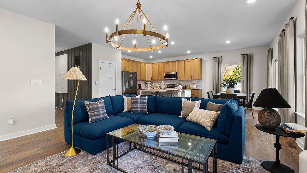 Stylish open-concept living room with a blue sectional sofa, modern chandelier, and adjacent kitchen featuring wooden cabinets and stainless steel appliances.