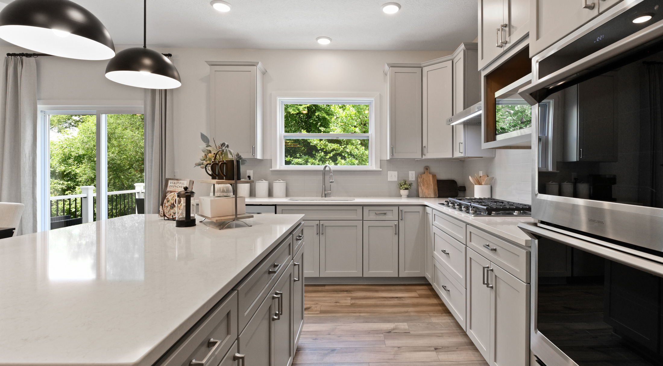 Modern kitchen with gray cabinetry, white countertops, stainless steel appliances, and large windows overlooking greenery.