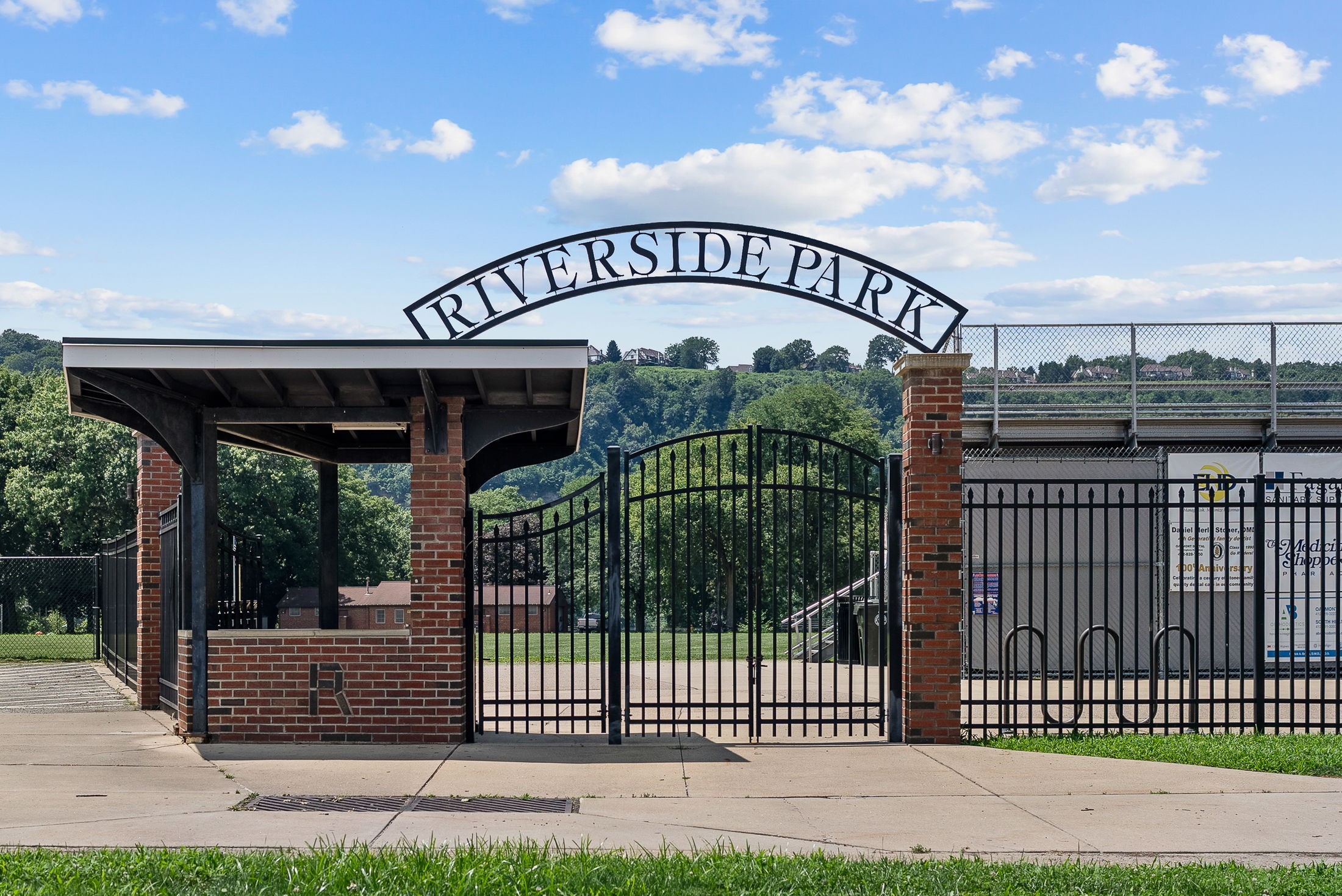Entrance gate of Riverside Park with brick pillars and metal arch under a blue sky.