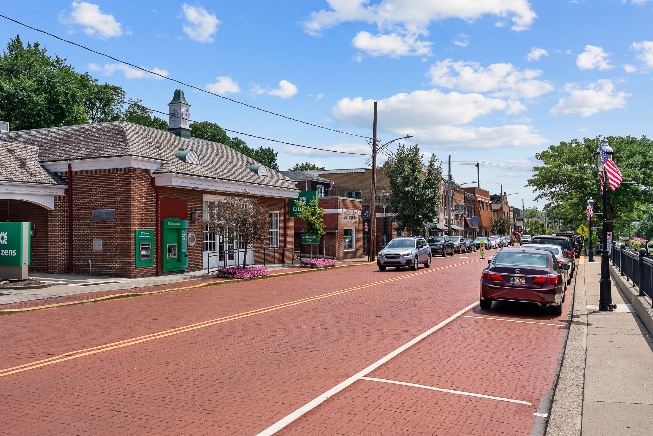 A charming small-town street scene featuring a brick building with ATM machines, parked cars, and an American flag-lined sidewalk under a blue sky.