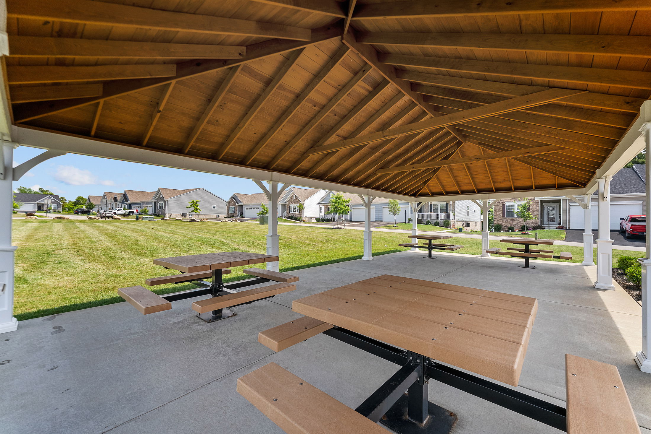 A spacious wooden pavilion with picnic tables overlooks a well-manicured lawn in a suburban neighborhood.