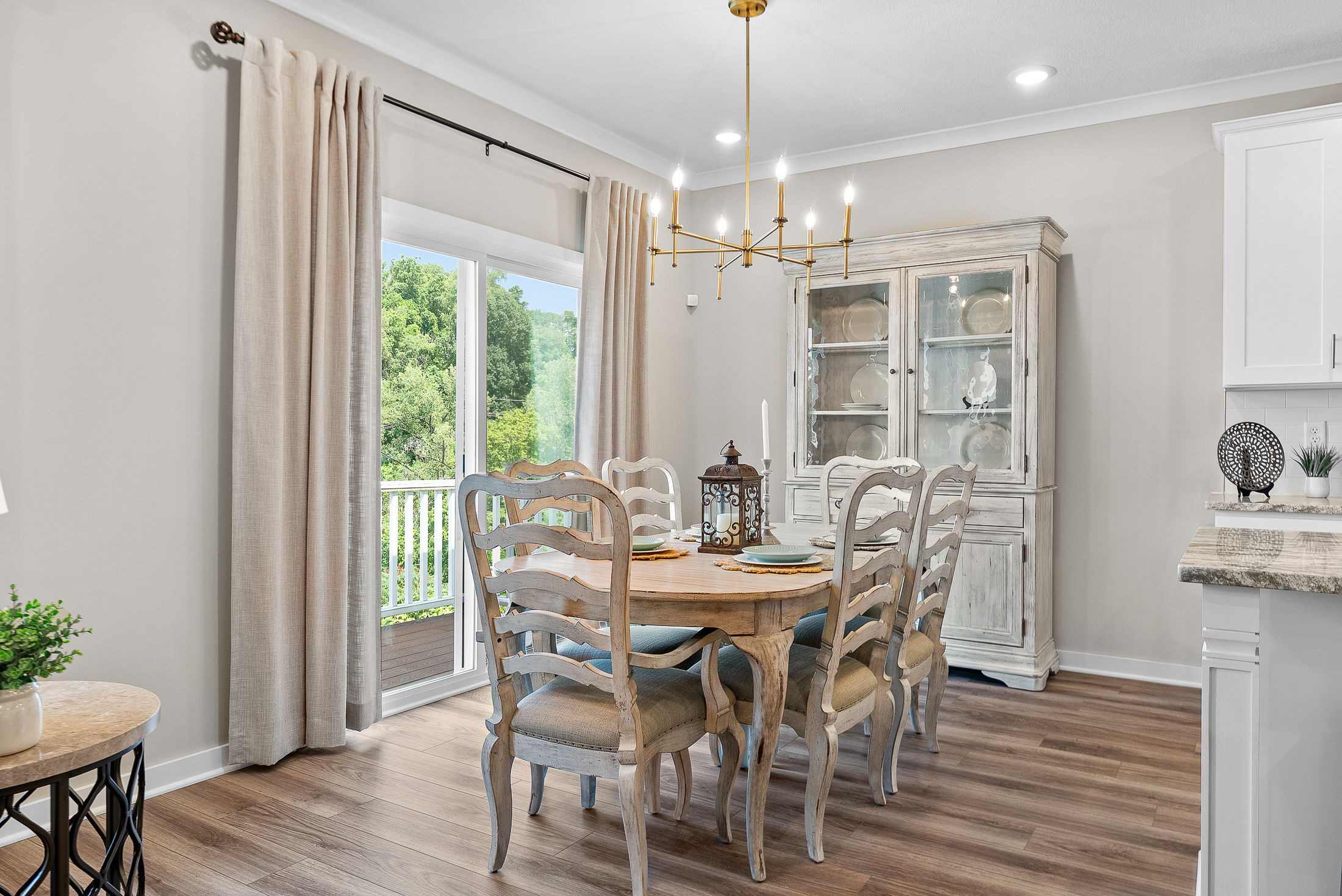 Elegant dining room with wooden table, cushioned chairs, a chandelier, and large bright windows showcasing a lush green view.