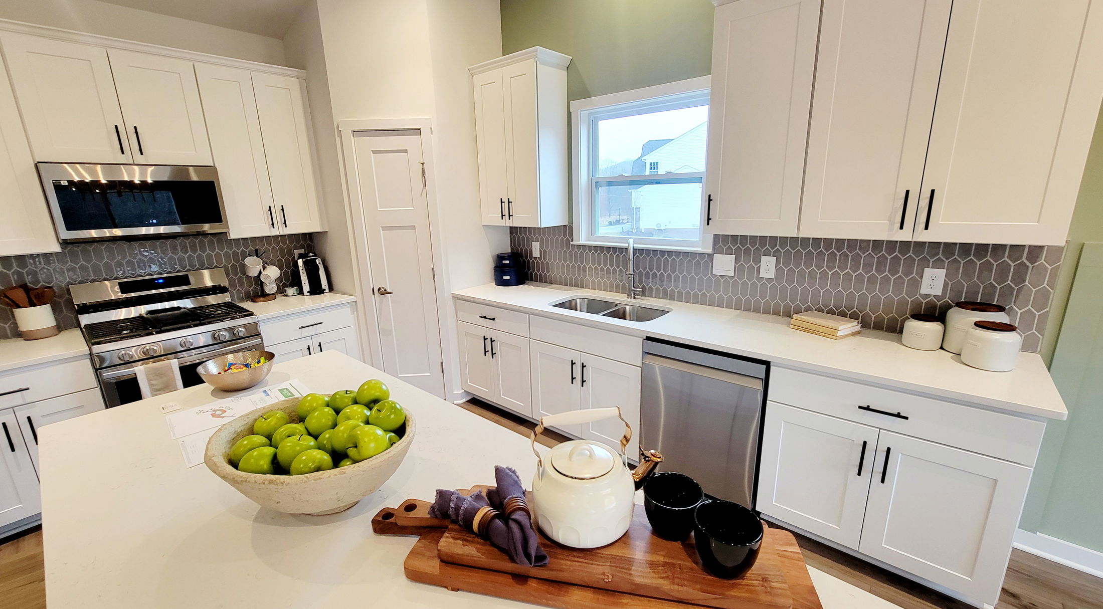 Modern kitchen with white cabinets, stainless steel appliances, and a central island featuring a bowl of green apples and a teapot.