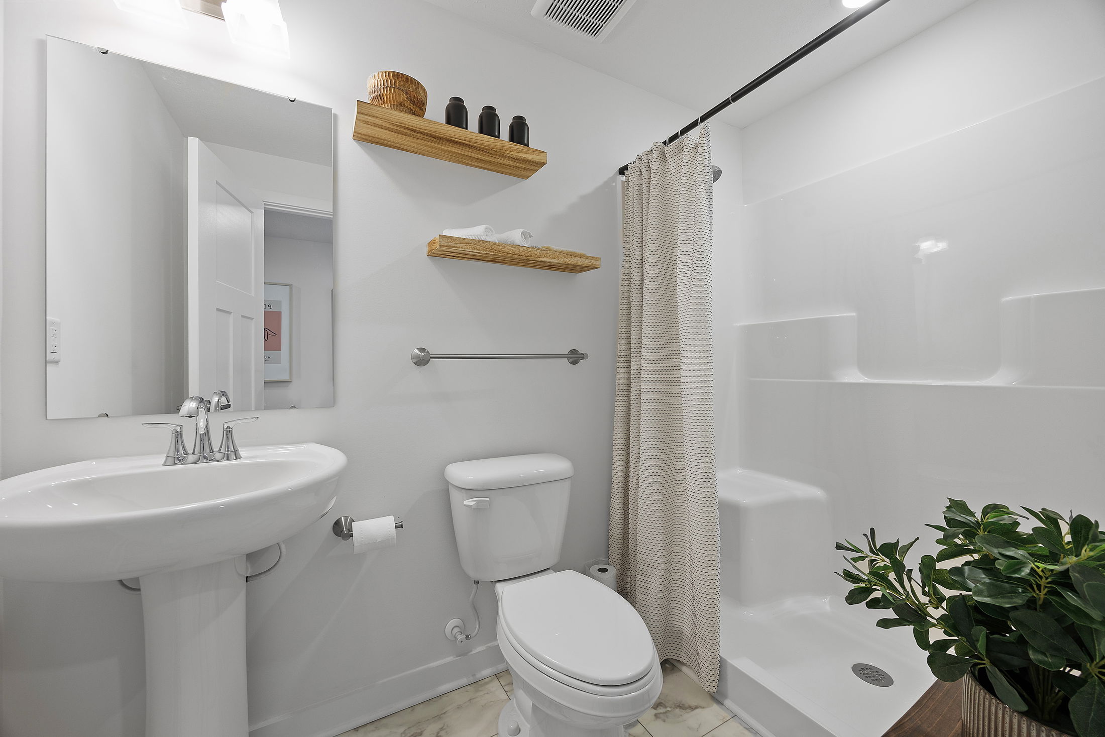A modern white bathroom with a pedestal sink, toilet, shower, and decorative wooden shelves featuring plants and toiletries.