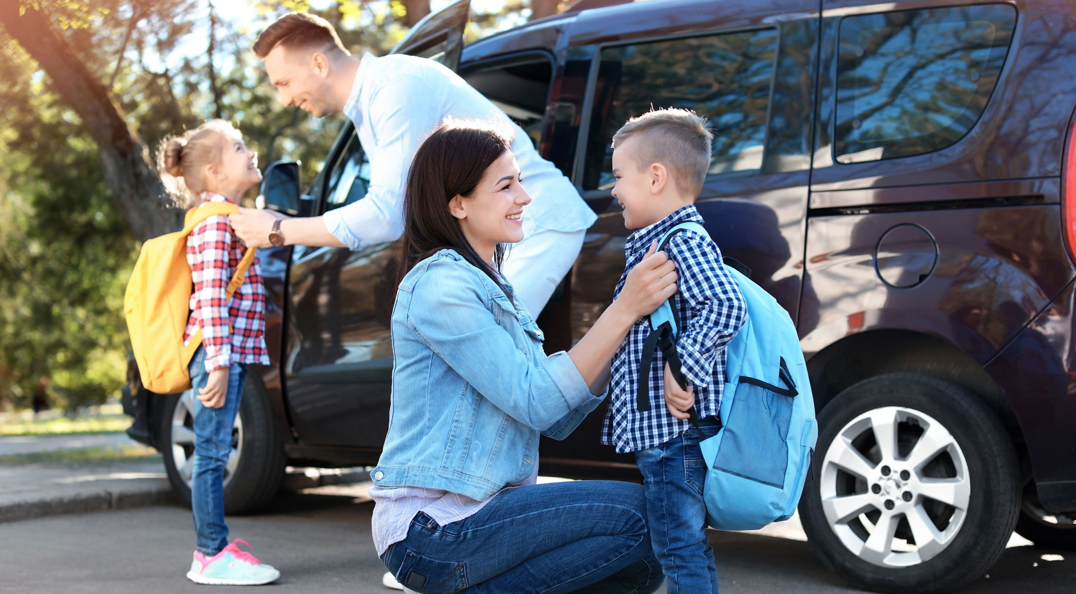 Parents dropping off children with backpacks at school near a parked car.