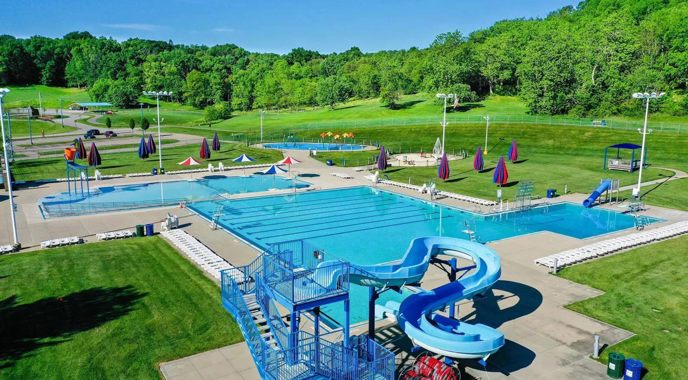 Aerial view of a vibrant outdoor water park featuring multiple swimming pools, a twisting blue water slide, and green landscaped surroundings.
