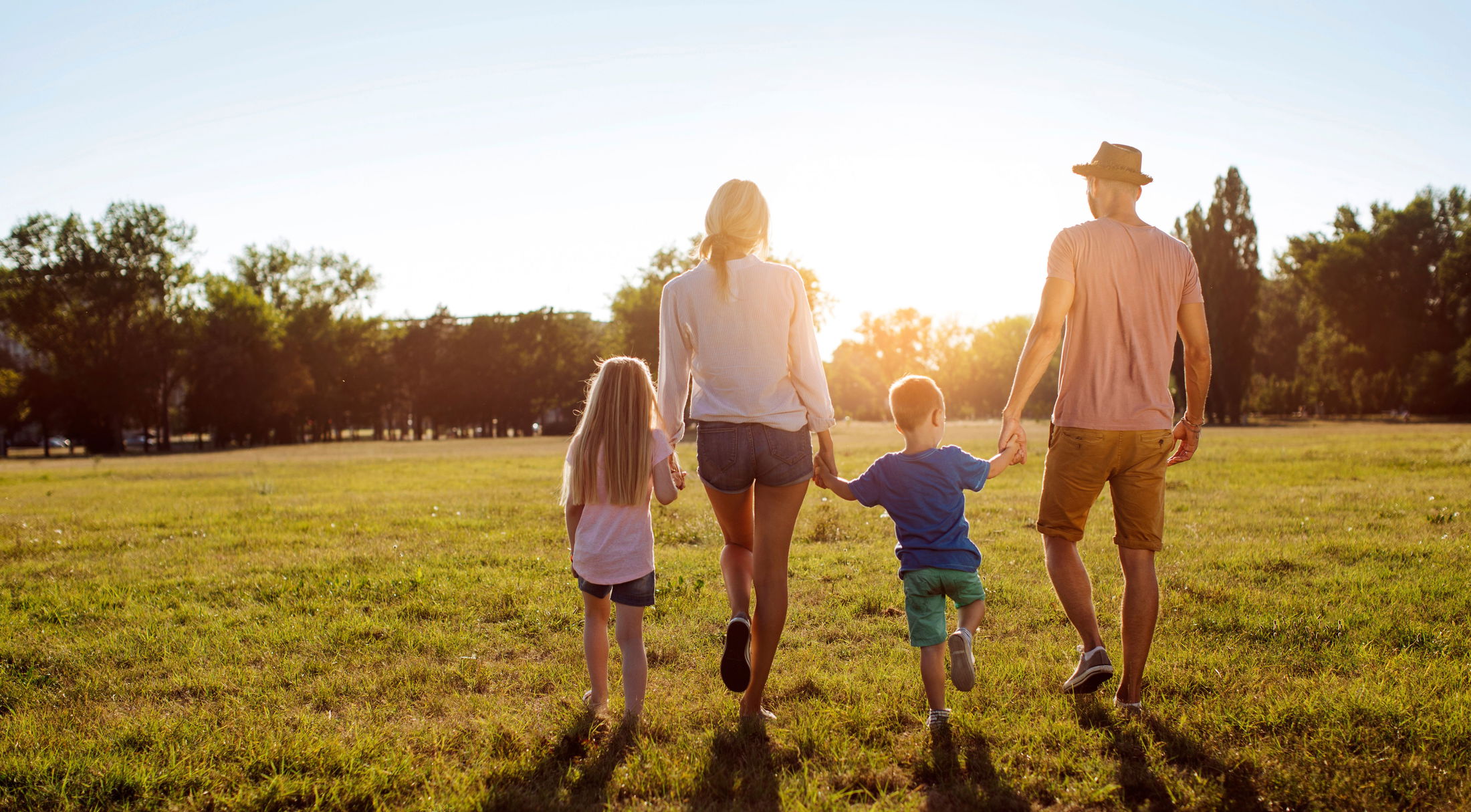 A family of four walking hand-in-hand in a sunny park during sunset, surrounded by lush greenery.