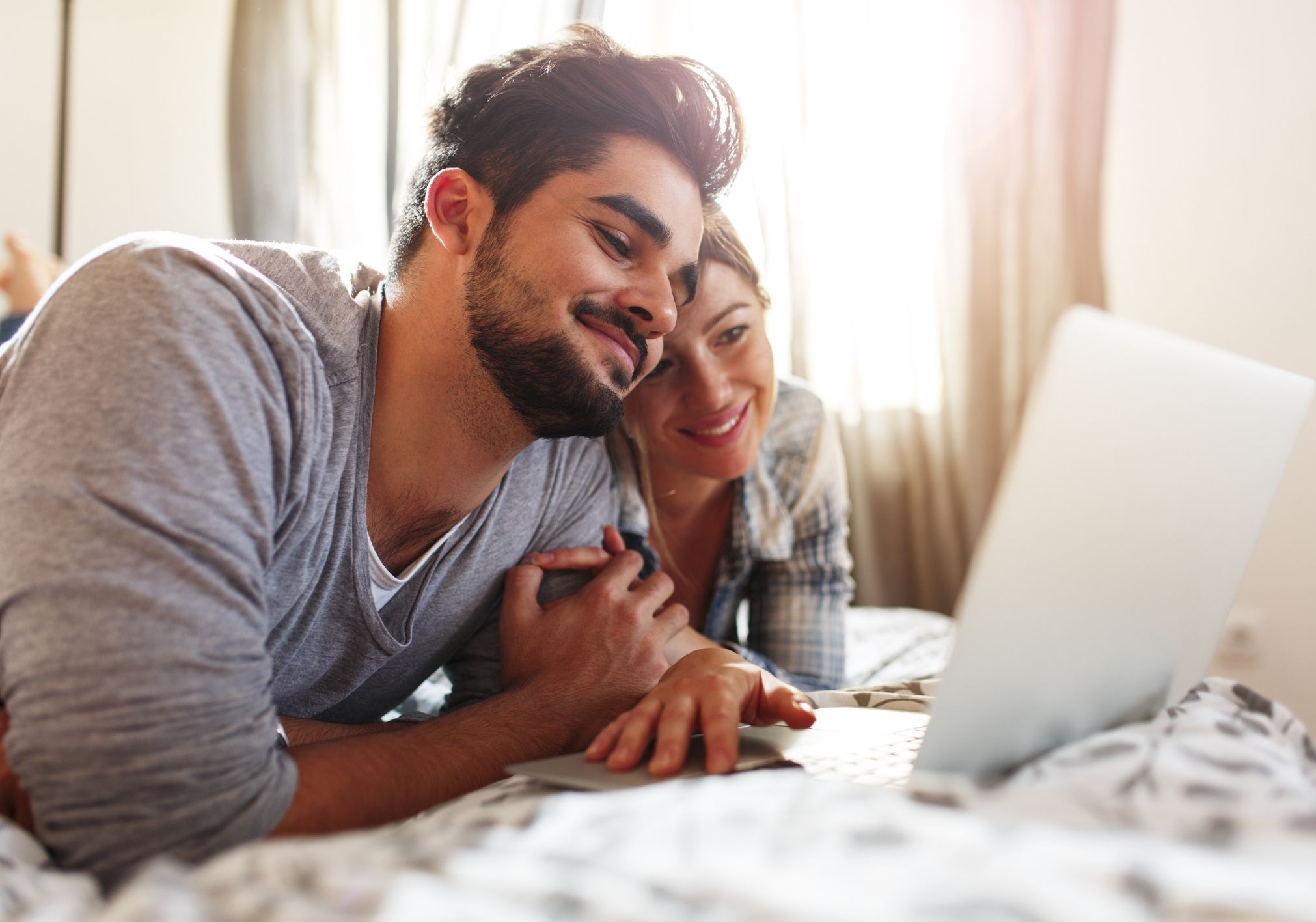 A couple lies on a bed, smiling while looking at a laptop together.