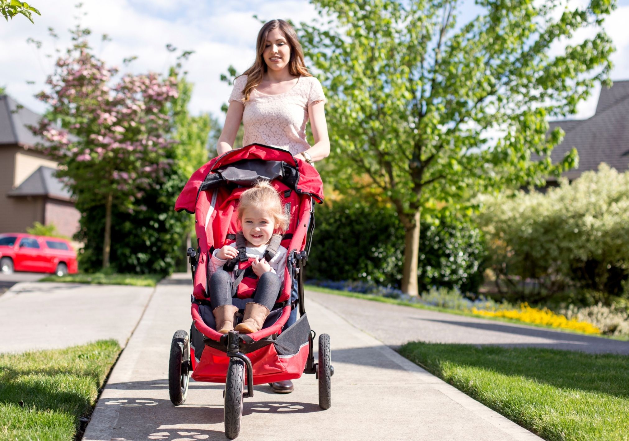 A woman pushes a child in a red stroller along a paved pathway bordered by greenery and blooming flowers.