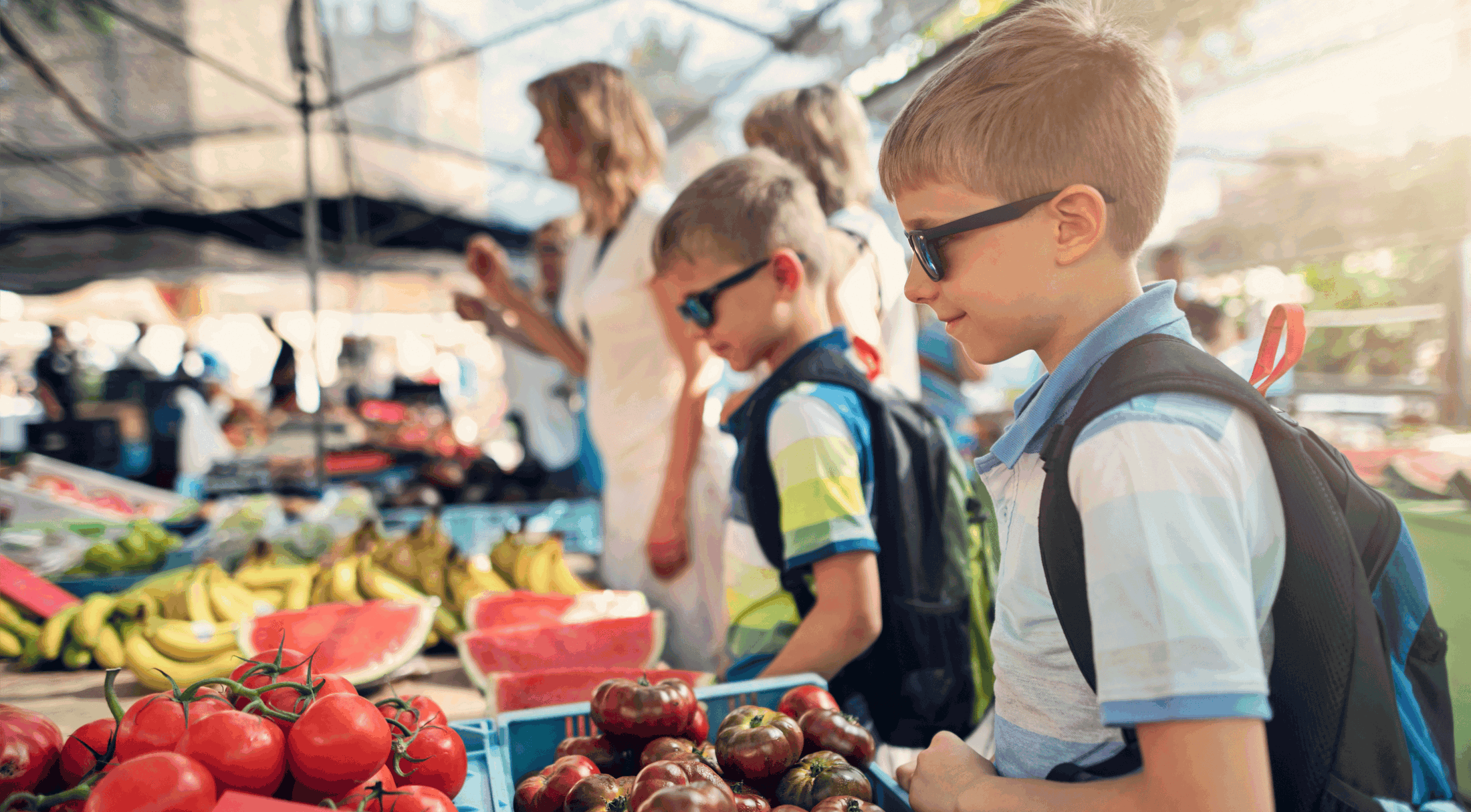 Two children with backpacks and sunglasses explore a lively outdoor farmers market filled with fresh produce like tomatoes, bananas, and watermelon.