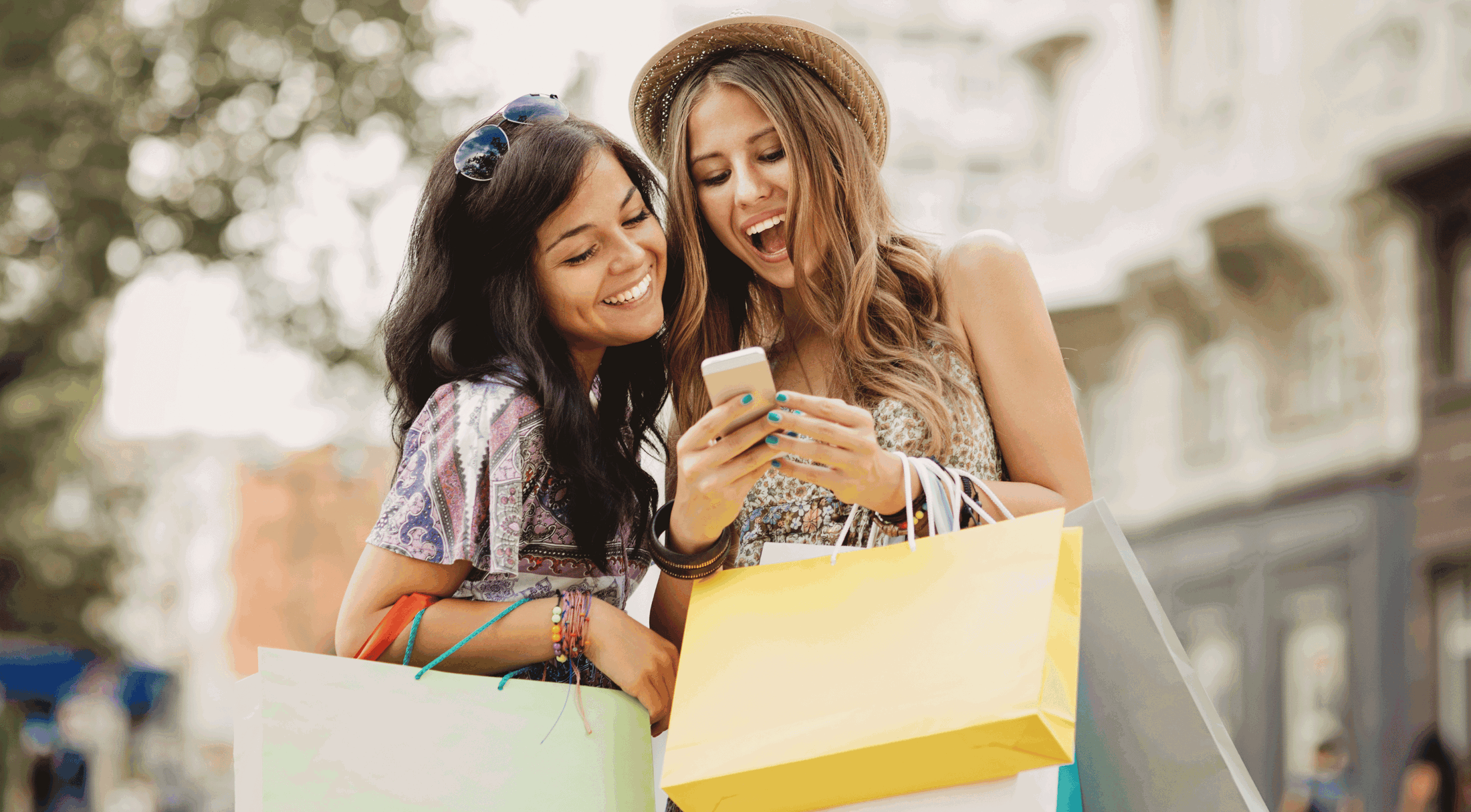 Two young women with shopping bags smiling and looking at a smartphone outdoors, enjoying a day of shopping.