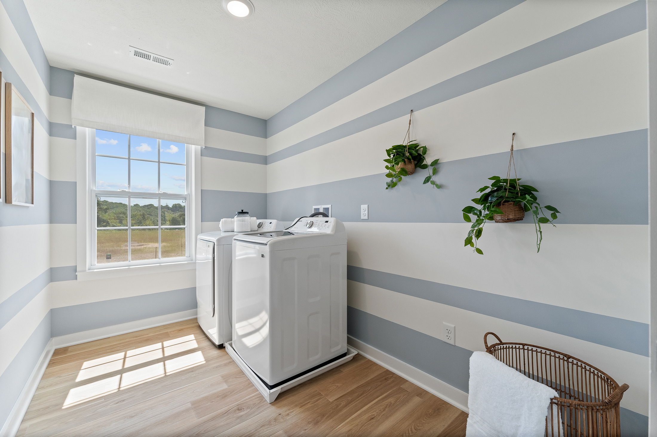Laundry room with striped blue and white walls, featuring a washer, dryer, hanging plants, and a large window.
