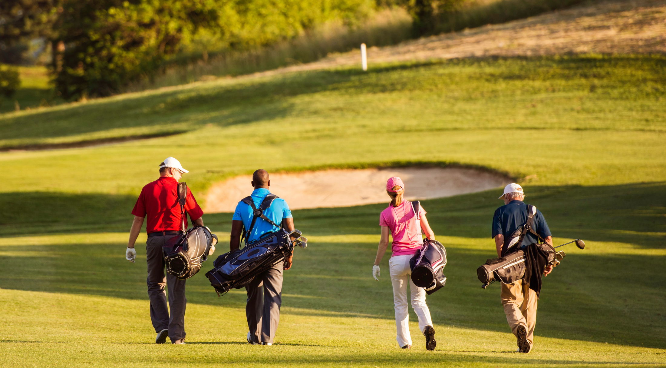 Four golfers walking on a lush golf course carrying bags near a sand bunker.