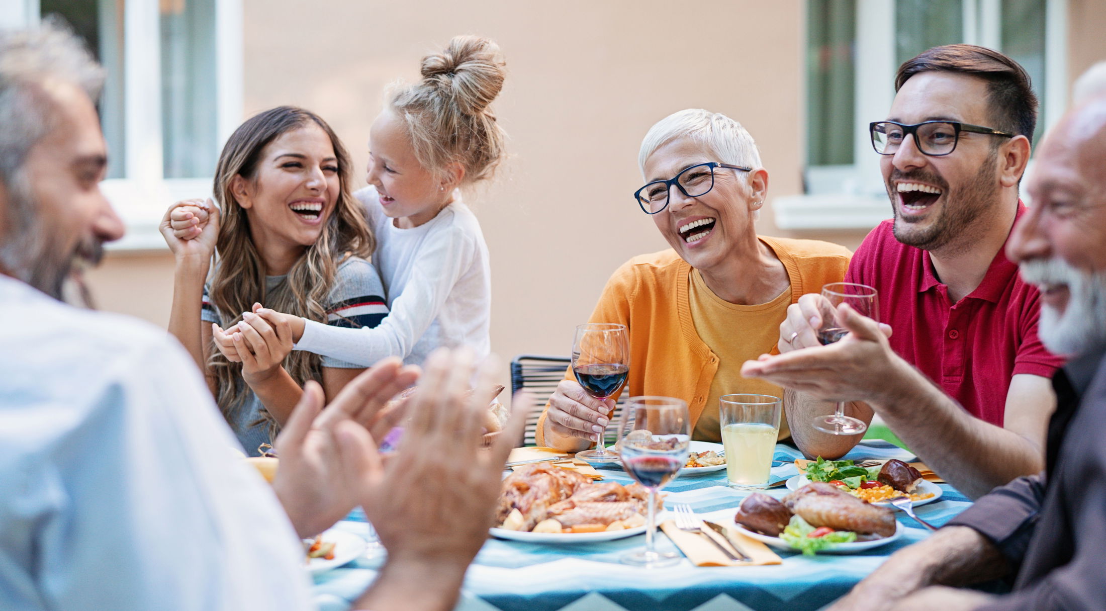 A diverse group of smiling people enjoying a joyful outdoor family meal together, sharing laughter and wine around a table filled with food.