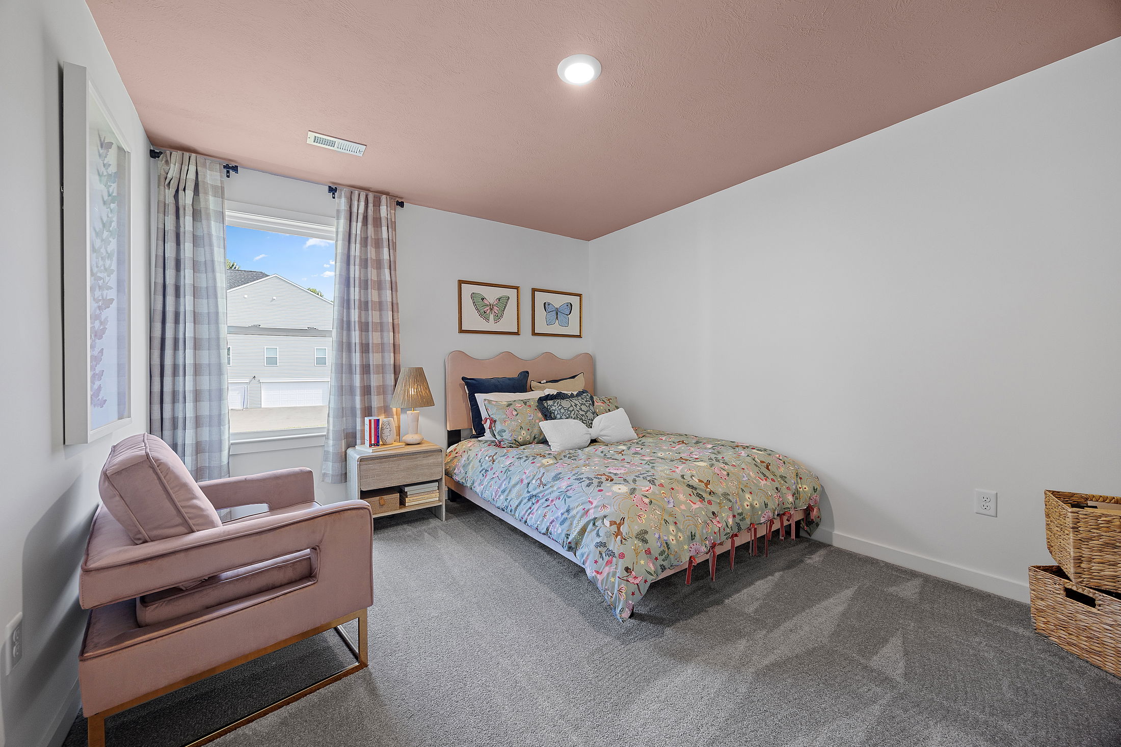 Cozy bedroom featuring a floral bedspread, pink armchair, and butterfly wall art, with checkered curtains framing a bright window.