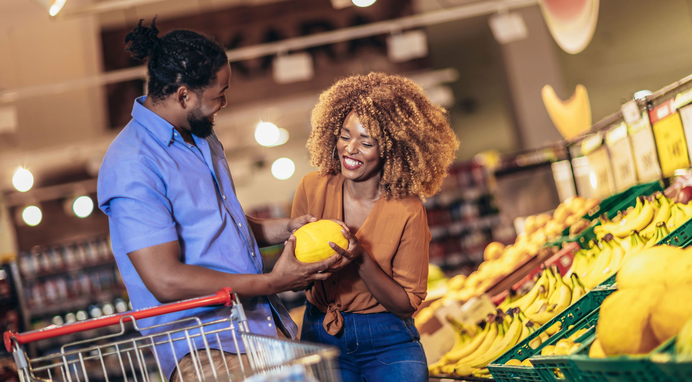 A smiling couple shopping for fresh produce in a grocery store, holding a yellow melon near a display of bananas.