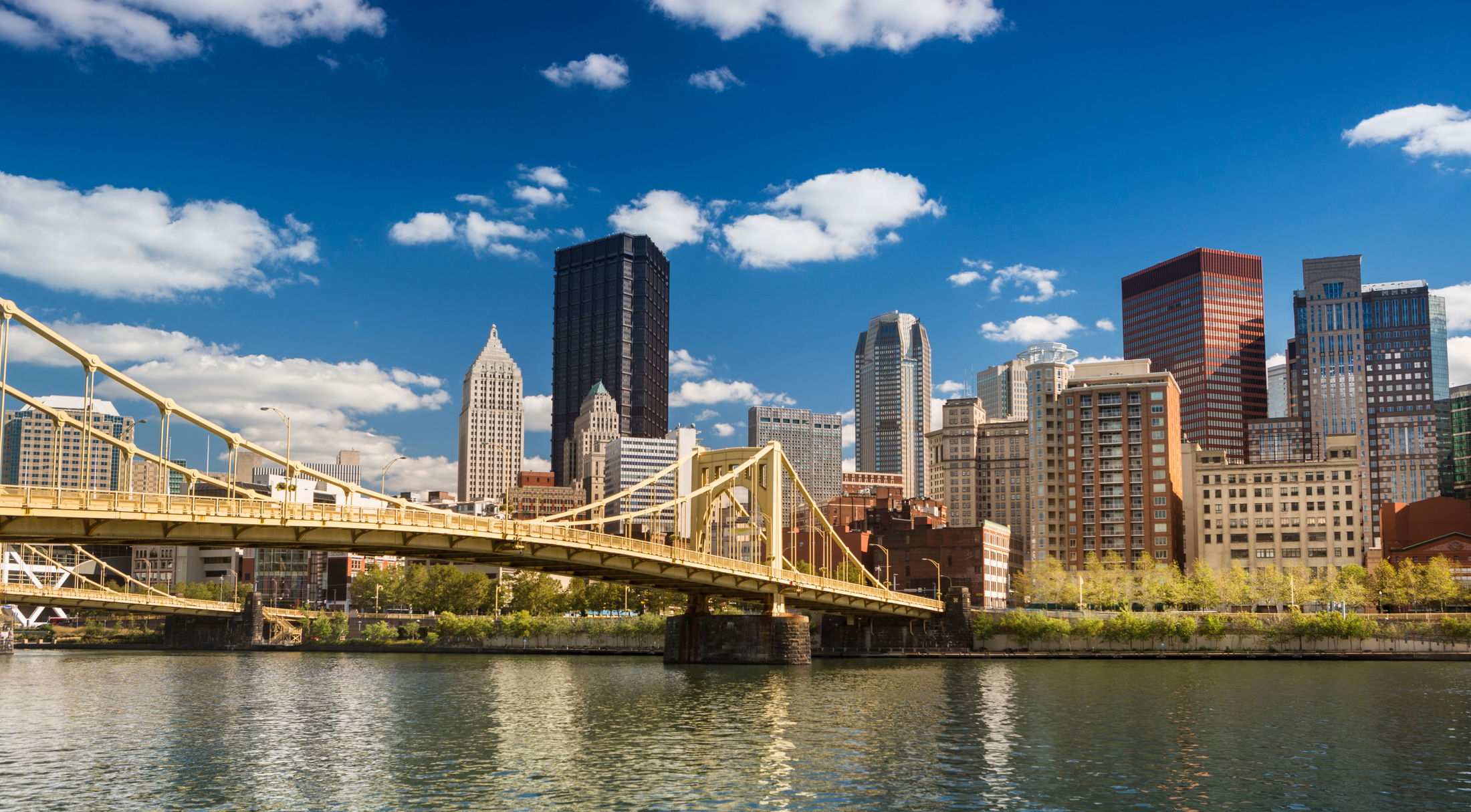 Scenic view of Pittsburgh skyline with iconic yellow bridge reflecting in the river under a blue sky.