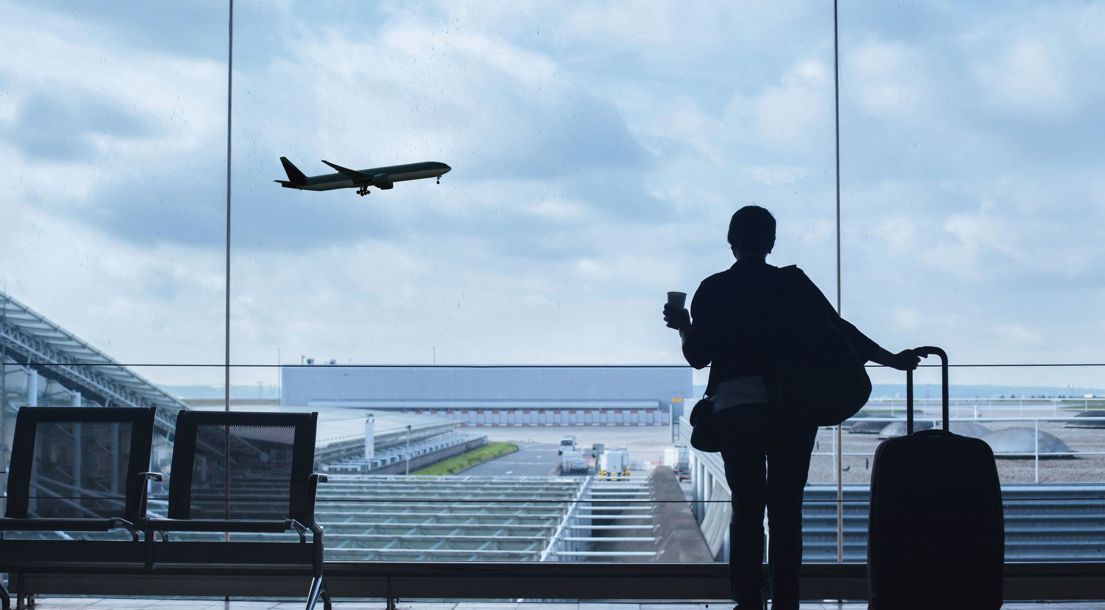 Silhouette of a traveler with luggage watching a plane take off through large airport windows.