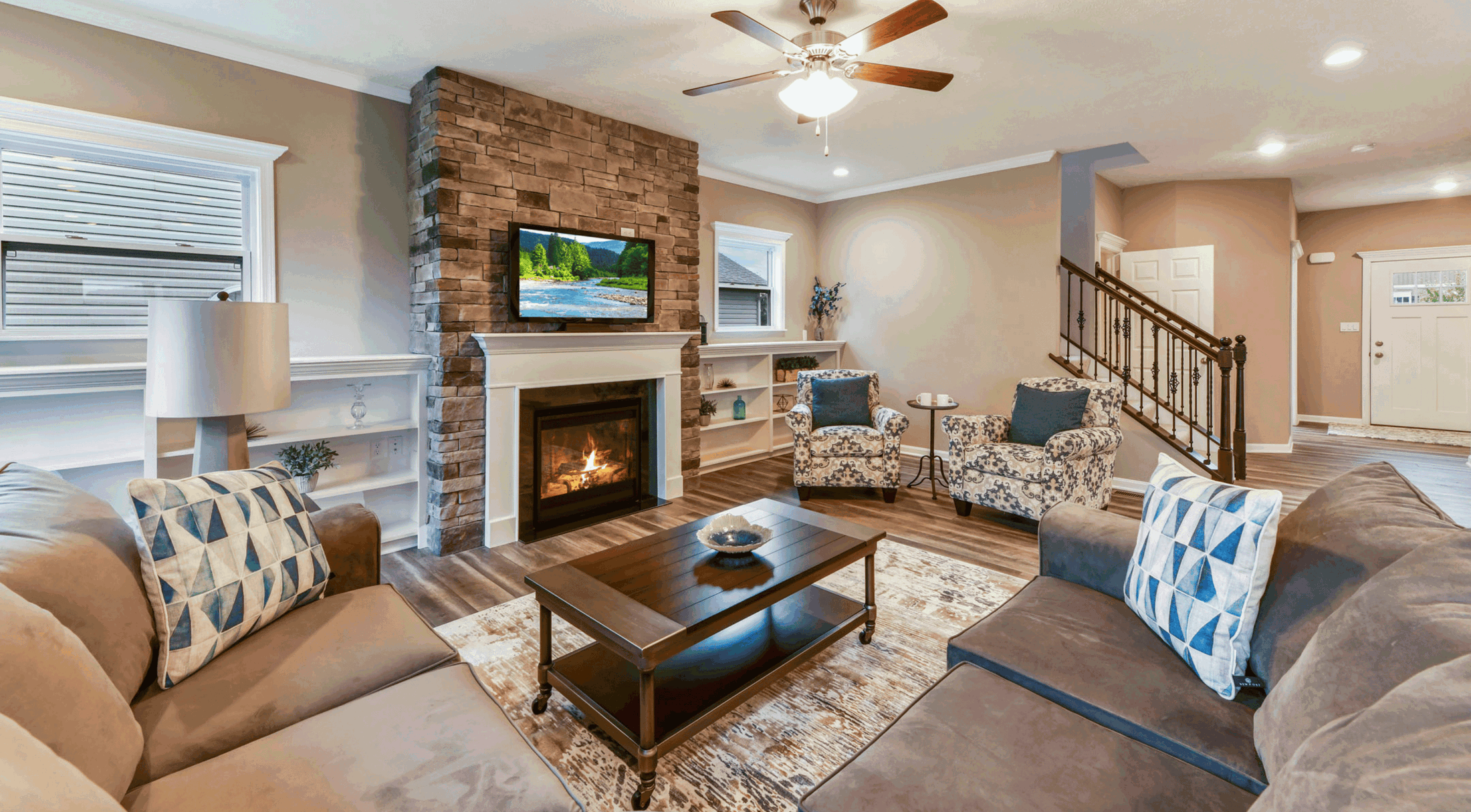 Modern living room with stone fireplace, wall-mounted TV, patterned armchairs, and brown sofas featuring blue geometric pillows.