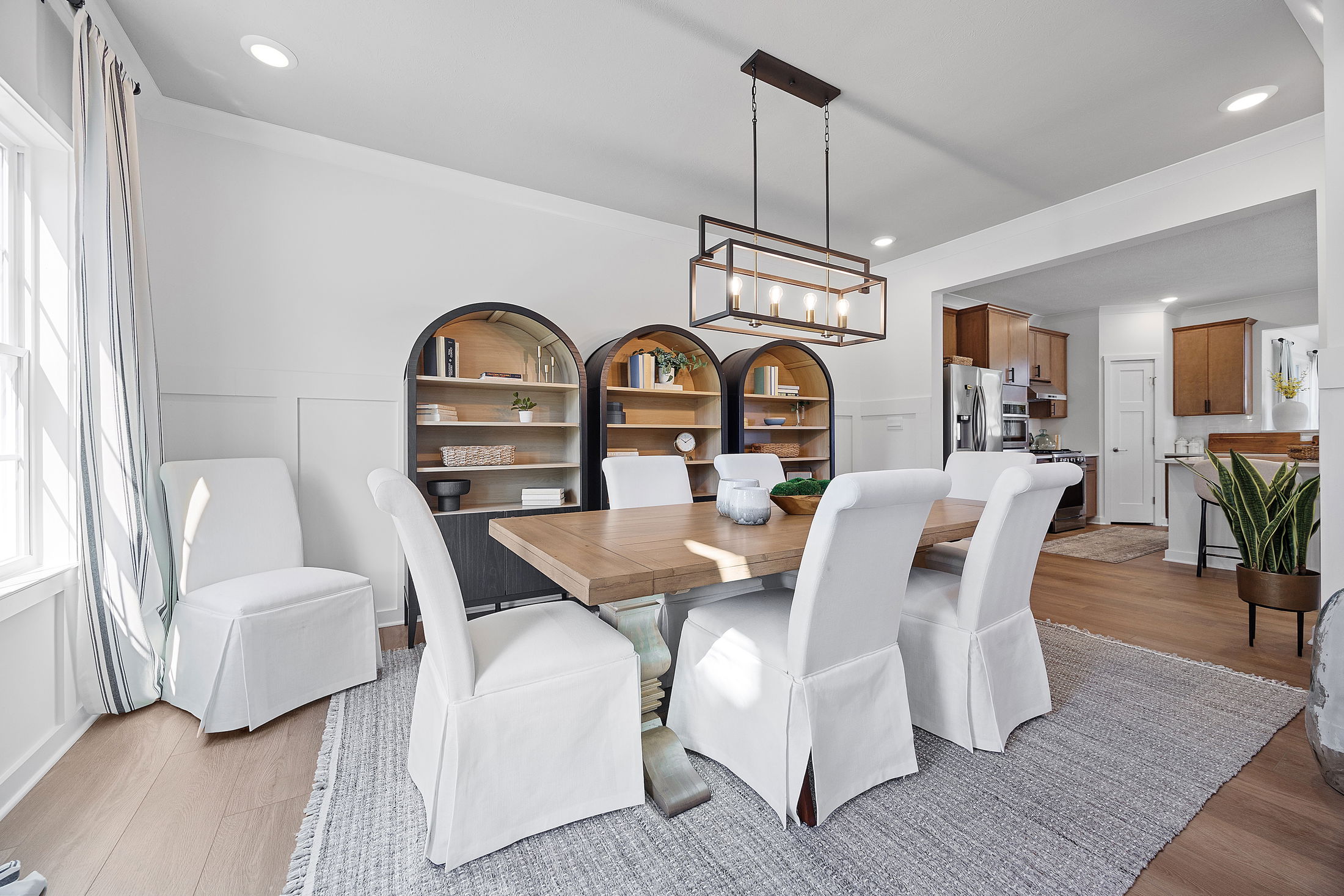 Modern dining room with a wooden table, six white slipcovered chairs, and stylish arched bookshelves, featuring a contemporary pendant light fixture.