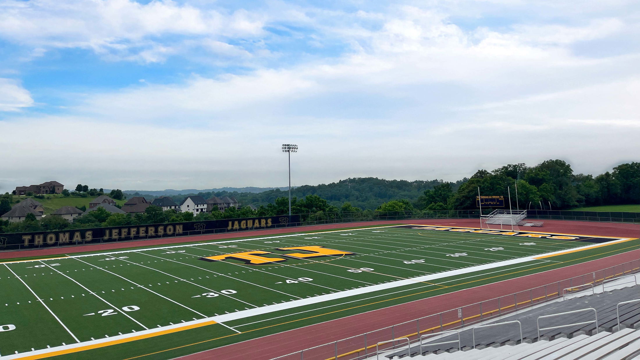 A panoramic view of Thomas Jefferson High School's football field and track, bordered by lush greenery and residential houses under a partly cloudy sky.