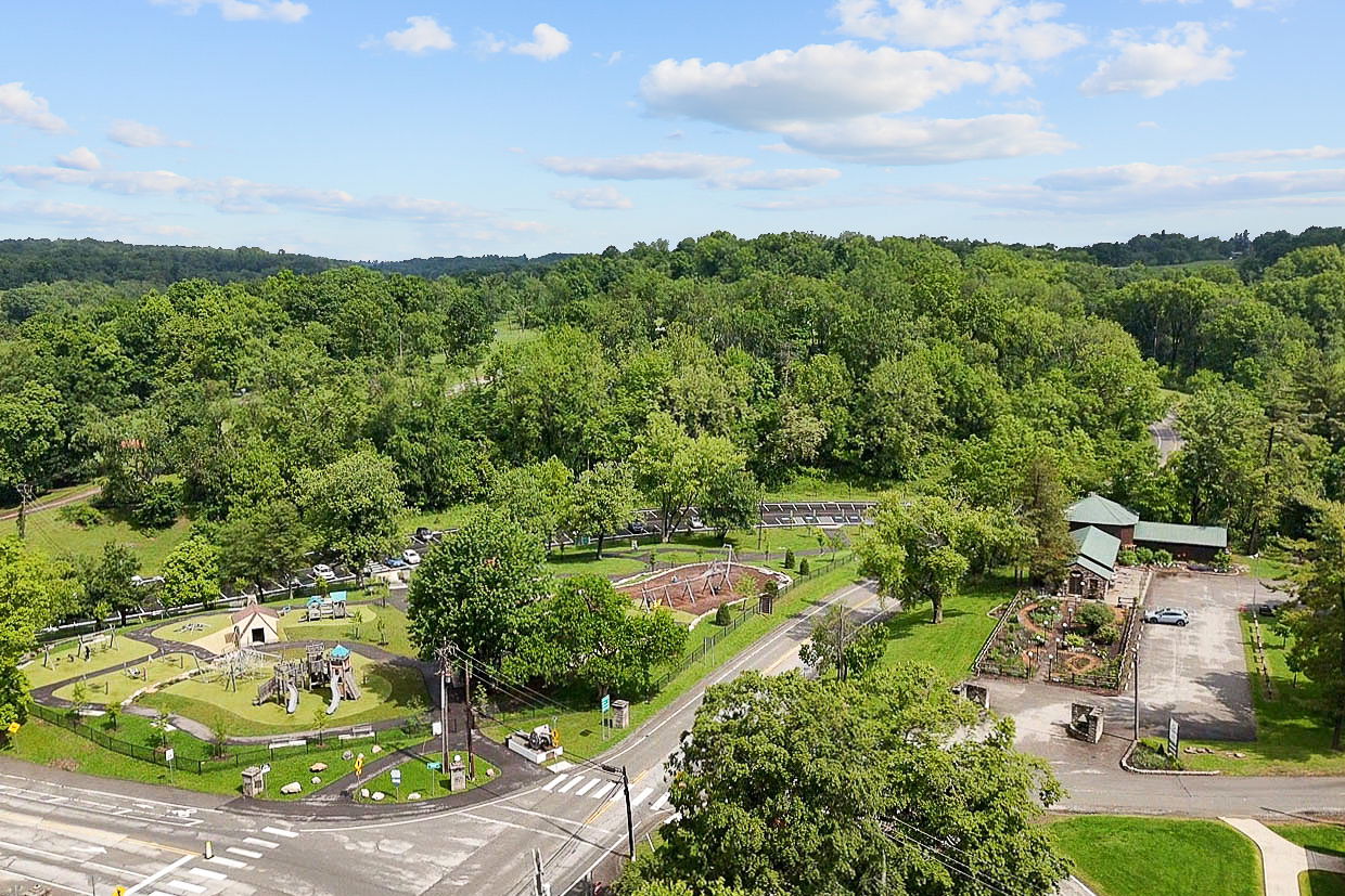 Aerial view of a lush green park featuring a well-maintained playground, walking paths, and surrounding wooded area under a partly cloudy sky.