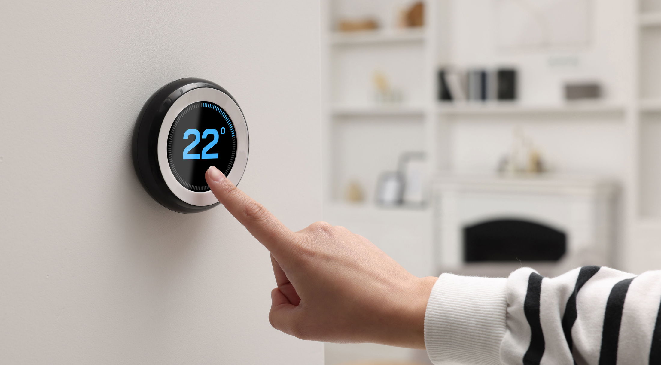 A person\\\\\\\\\\\\\\\'s hand adjusting a modern digital thermostat set at 22 degrees on a white wall.