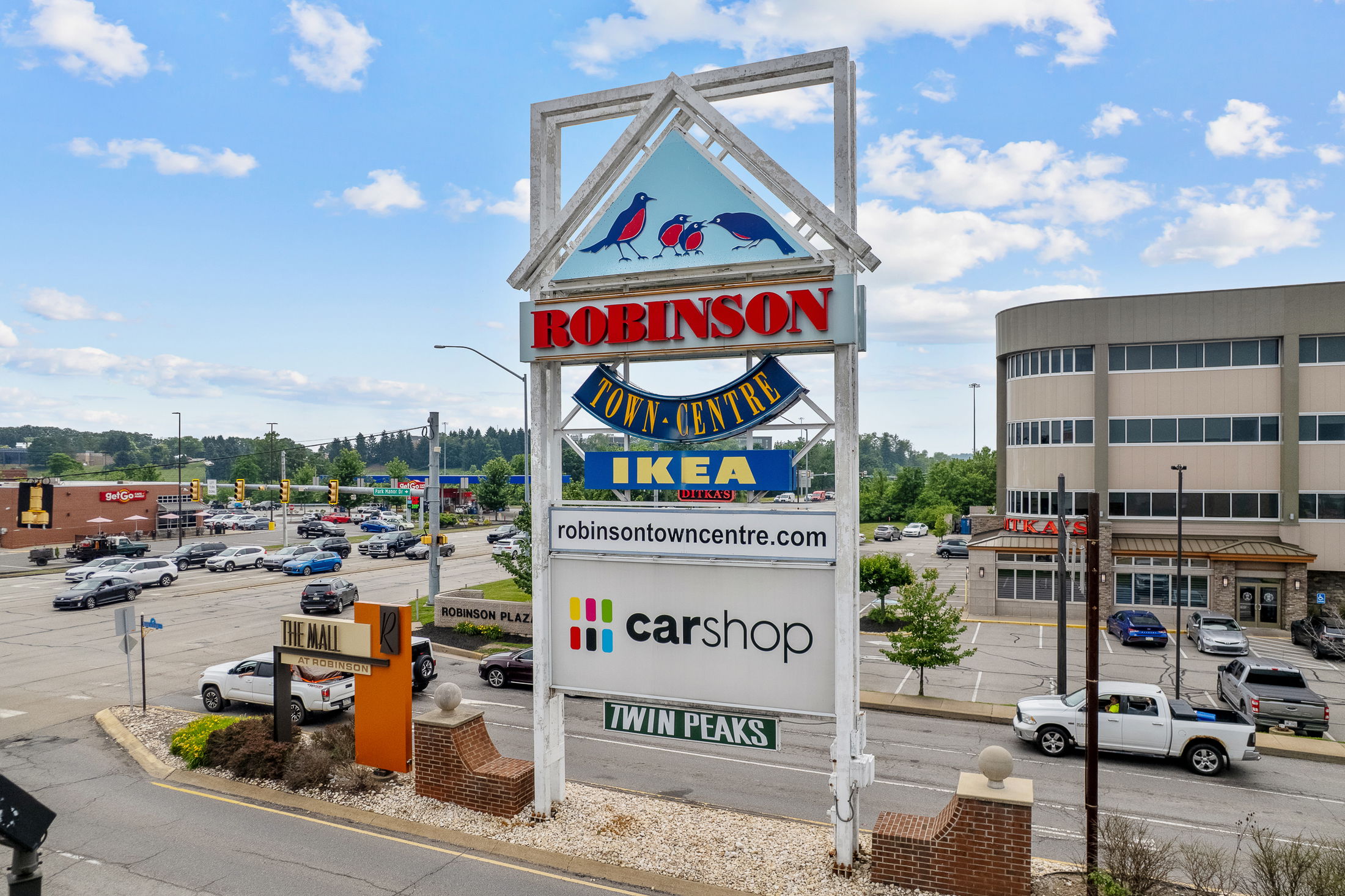 Large sign for Robinson Town Centre featuring IKEA, CarShop, and Twin Peaks, with nearby traffic and shops under a blue sky.