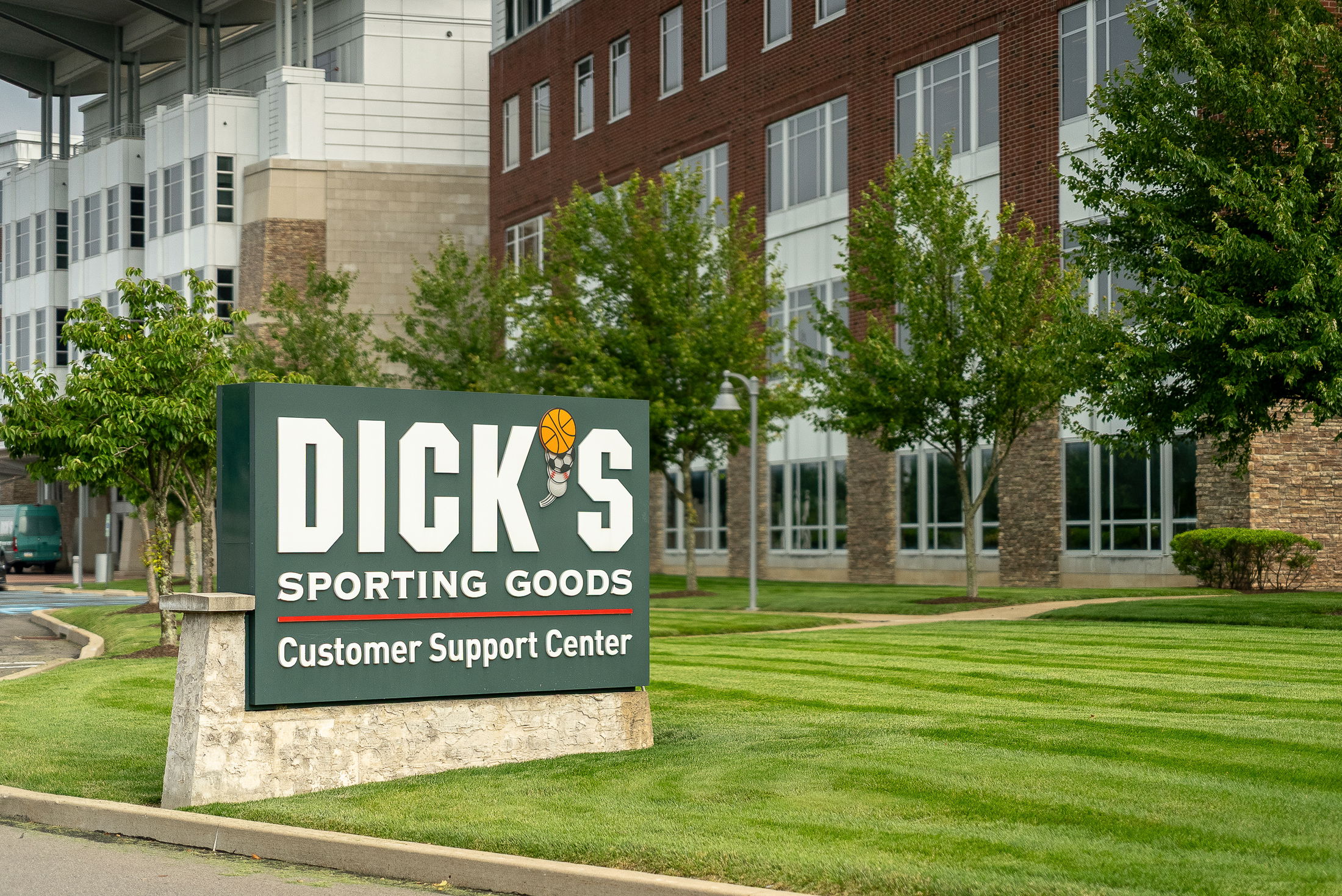 Front view of Dick\\\'s Sporting Goods Customer Support Center sign with lush green landscape and building in the background.