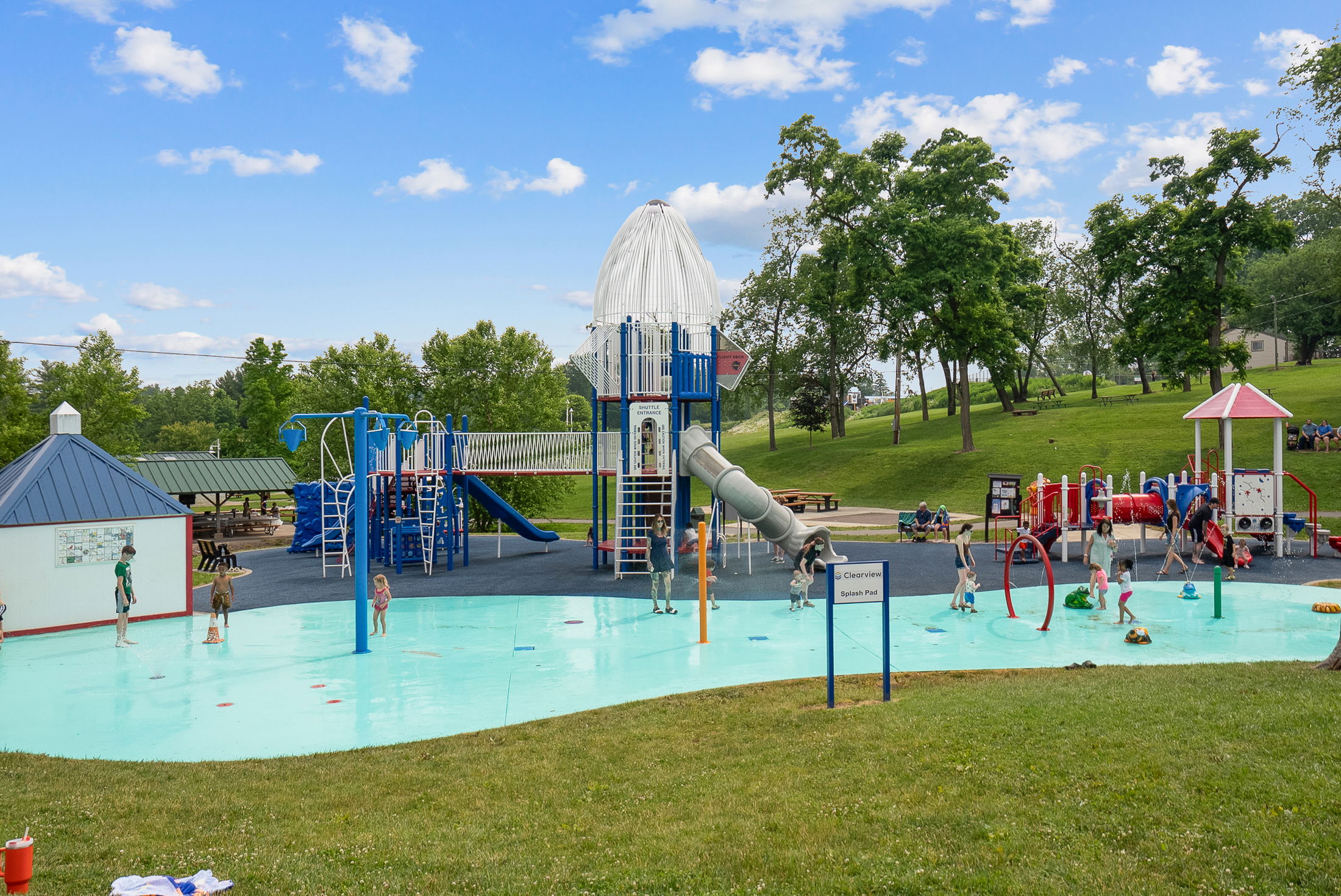 A vibrant children\\\'s playground with a rocket ship slide and splash pad surrounded by lush greenery under a blue sky.