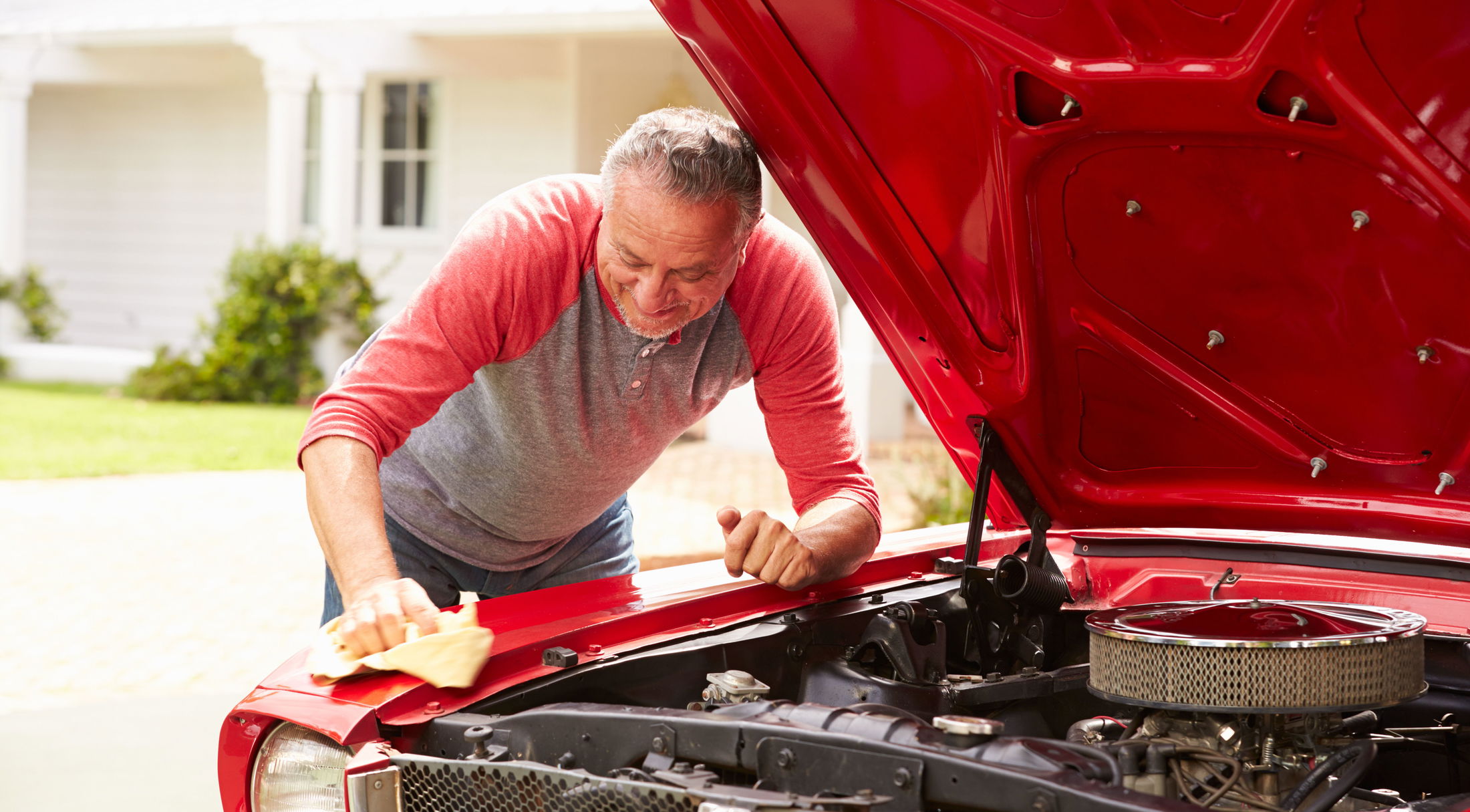 Man wearing a red and gray shirt cleaning the hood of a red car with an open engine.
