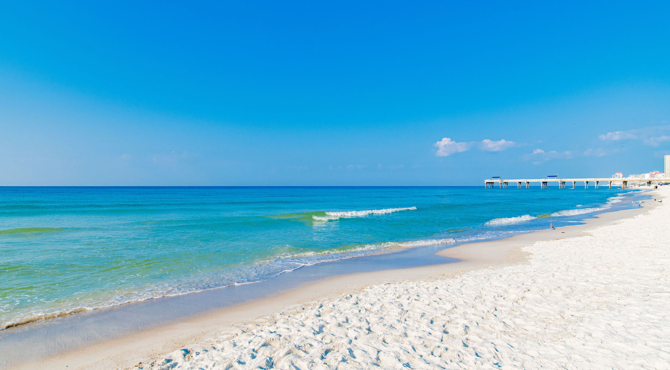 Sandy beach with turquoise ocean waves and a pier under a clear blue sky.