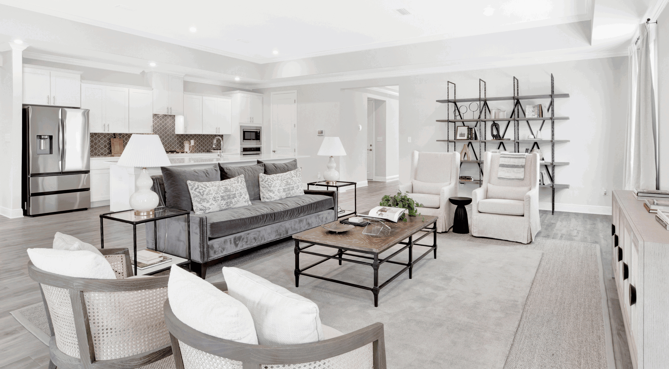Modern open-concept living room and kitchen with gray couch, stylish chairs, and stainless steel appliances.