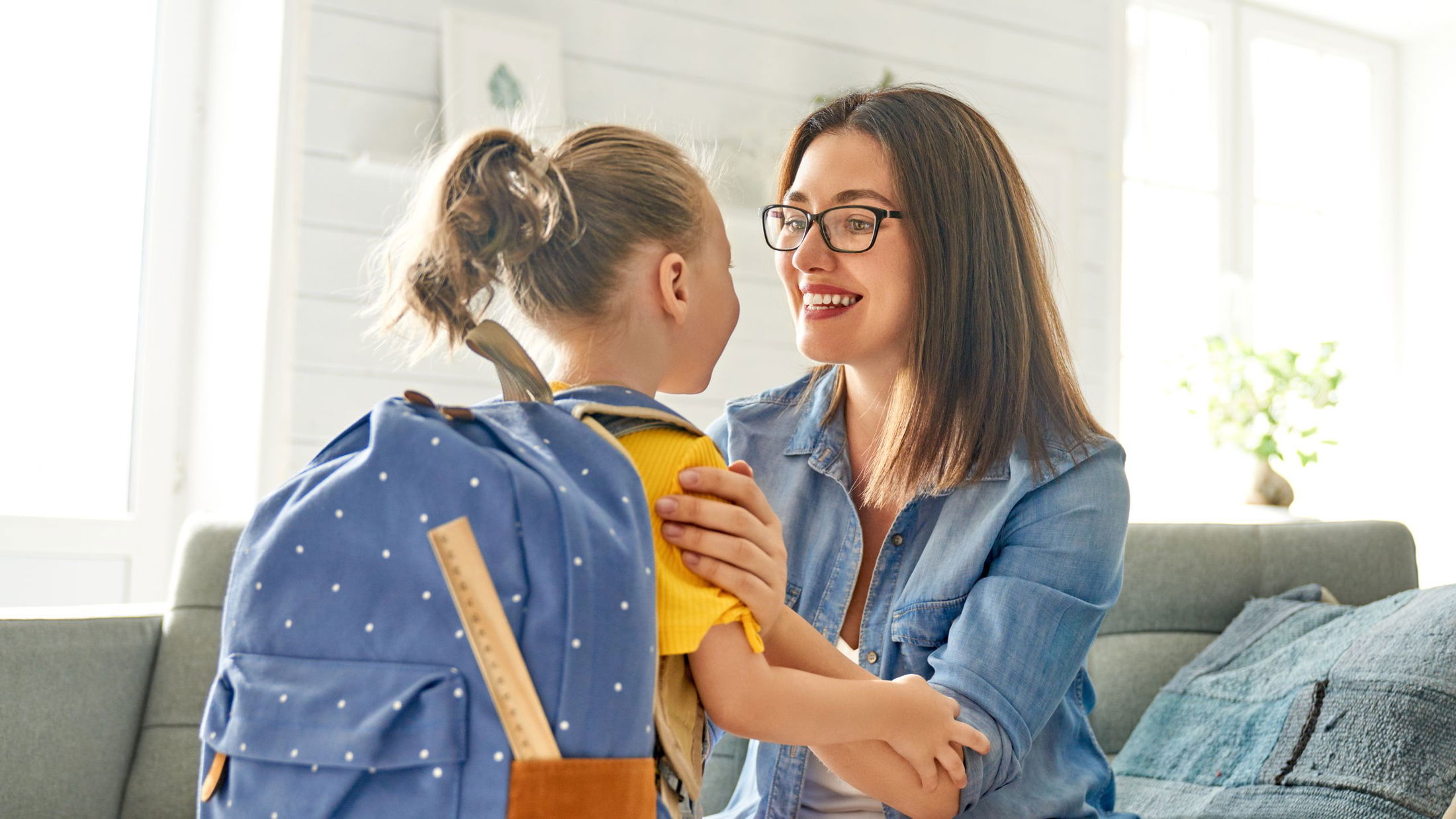 A mother smiles and encourages her daughter wearing a blue backpack before school in a bright living room.