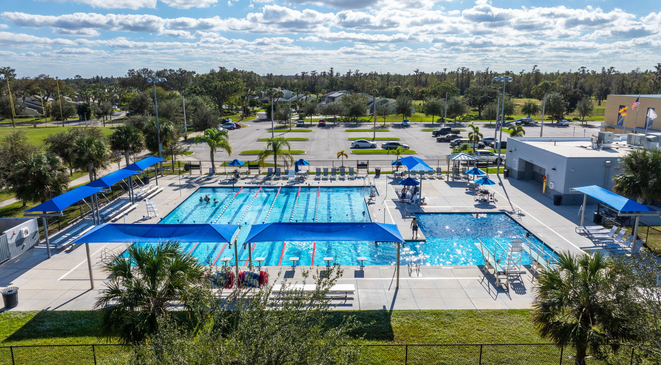 Aerial view of an outdoor swimming facility with multiple pools, blue shade structures, and landscaped surroundings under a partly cloudy sky.