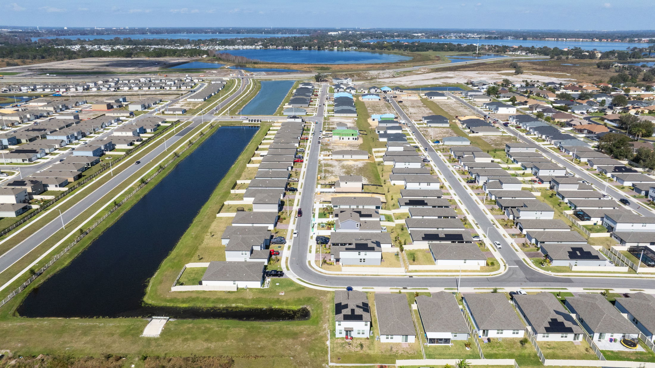 Aerial view of a modern suburban neighborhood with rows of houses, roads, and water canals, set against a backdrop of distant lakes.