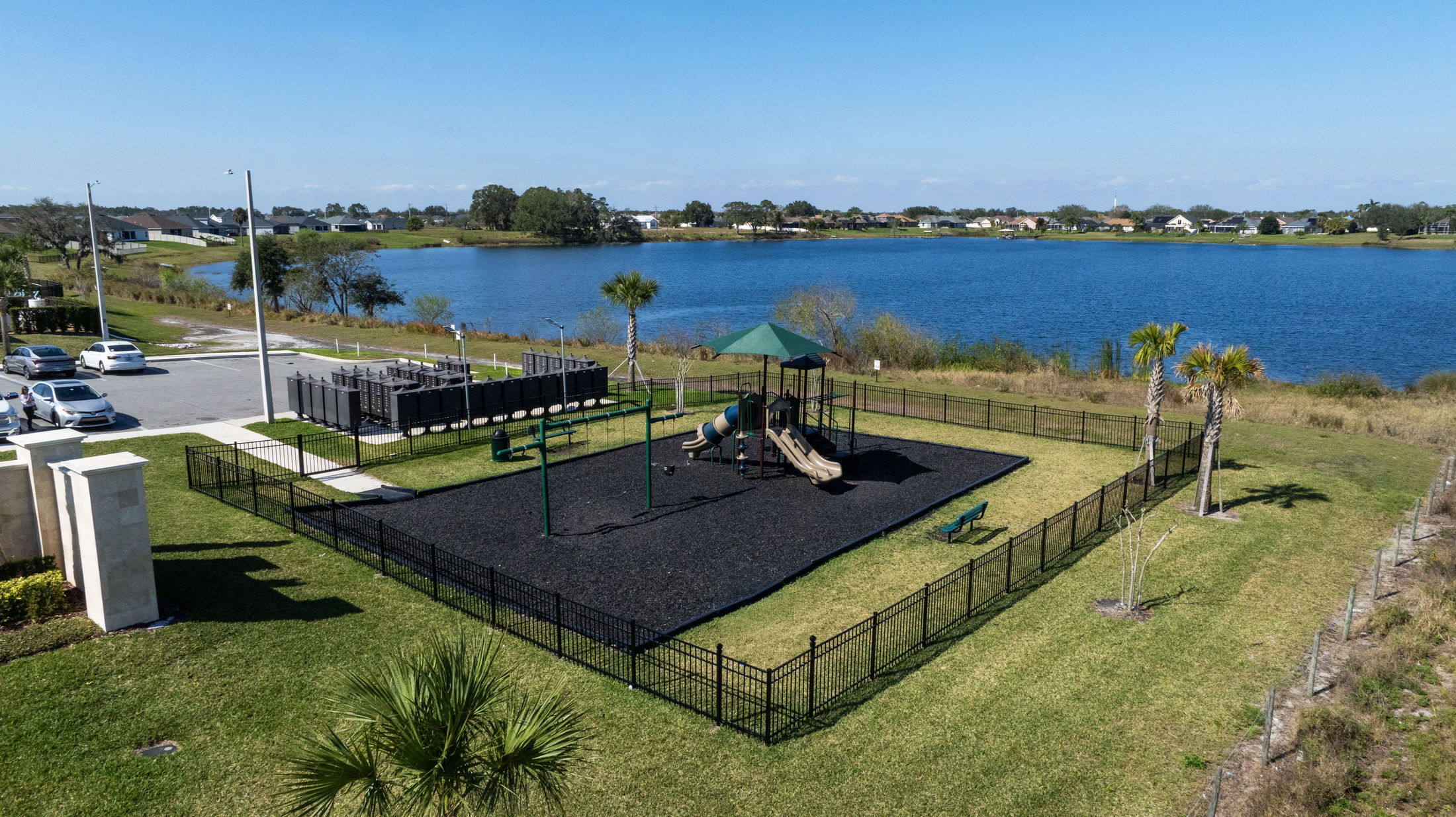 Aerial view of a fenced playground with swings and slides adjacent to a serene lakeside neighborhood, under a clear blue sky.