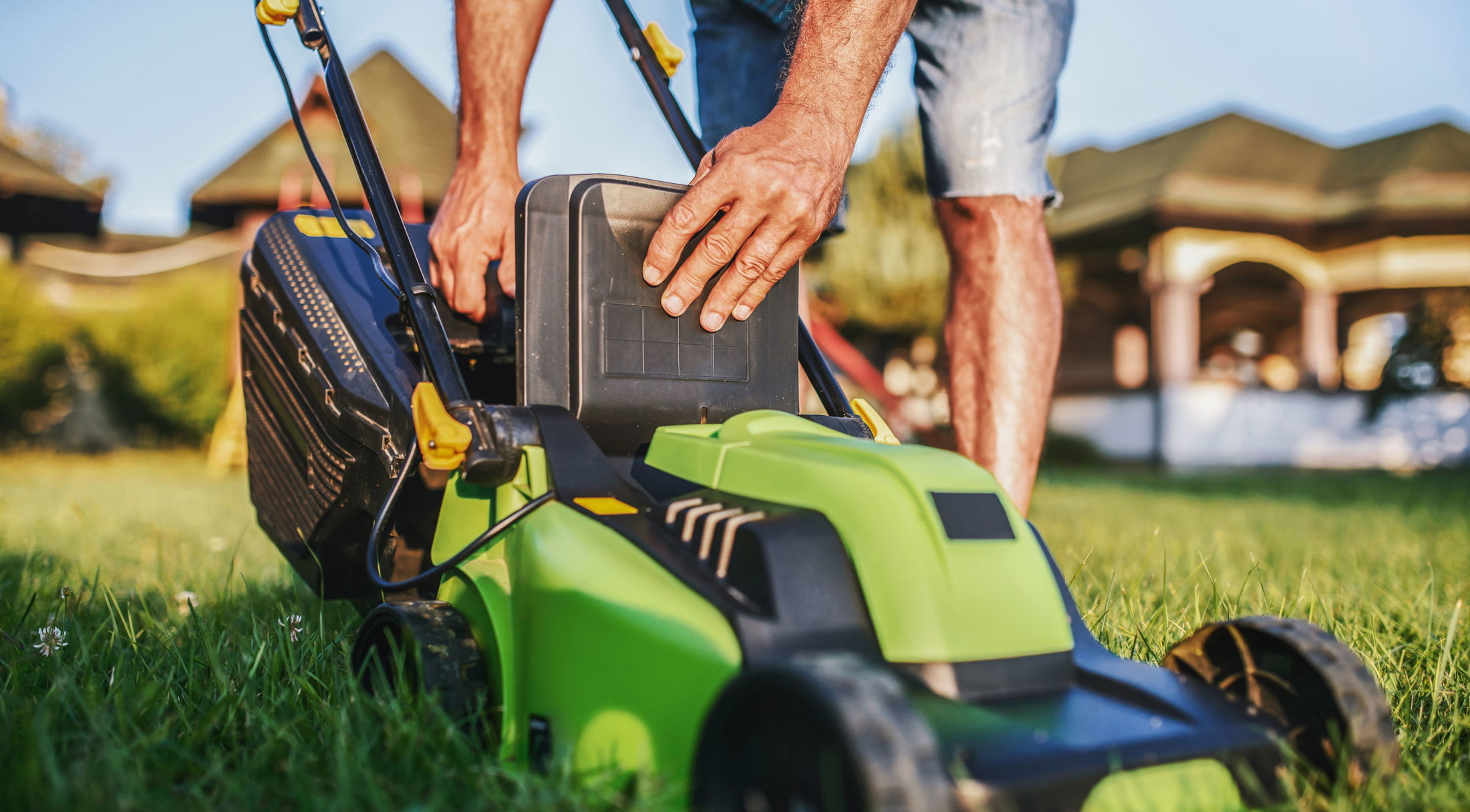 A person inserts a battery into a green electric lawn mower on a sunny day in a backyard garden.