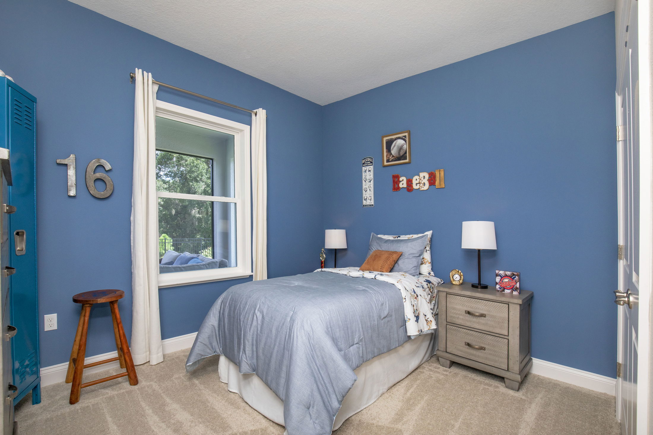 Blue-themed bedroom with baseball decor, featuring a single bed, wooden stool, and window with a view of greenery.