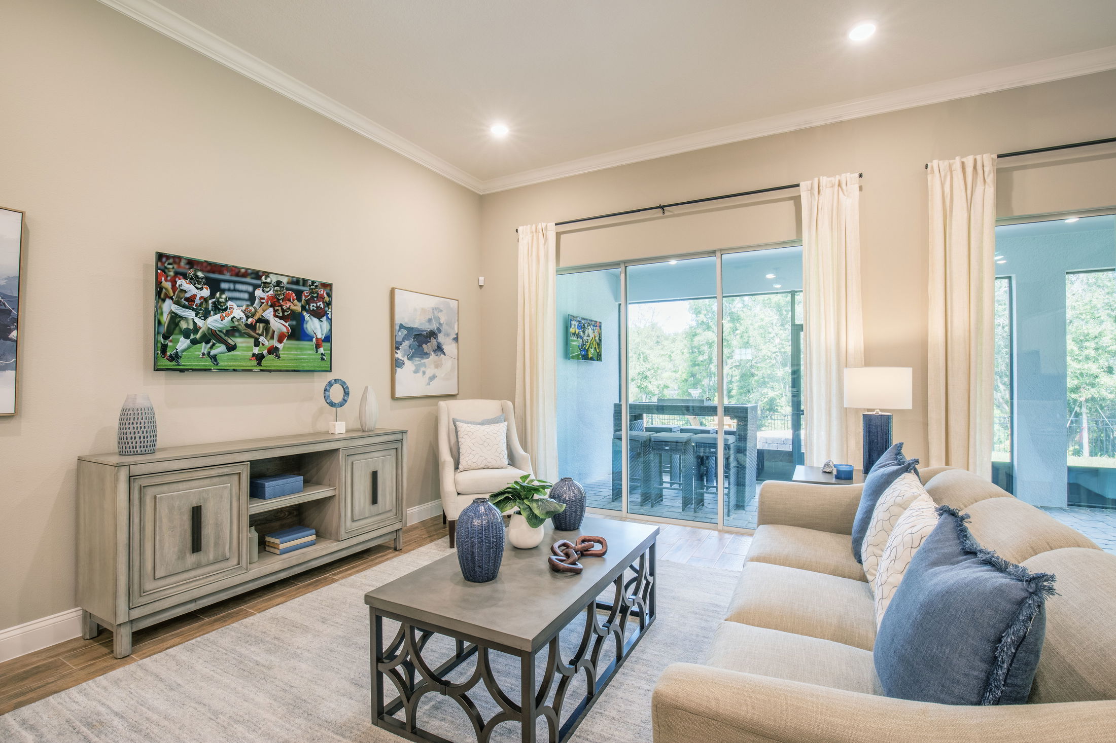 Modern living room with beige sofa, stylish coffee table, and a TV displaying a sports game, leading to a bright outdoor patio.