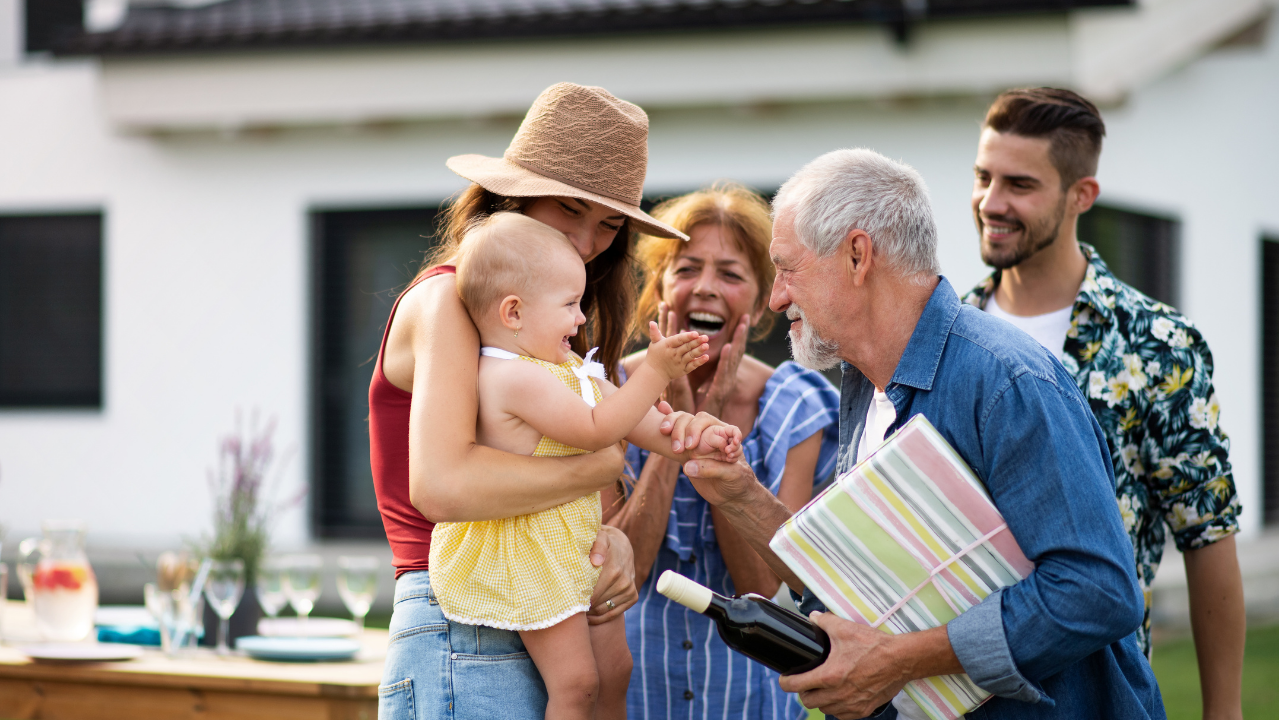 A family gathering in a backyard with four adults and a baby, where an elderly man is joyfully interacting with the baby while holding a gift and a wine bottle.
