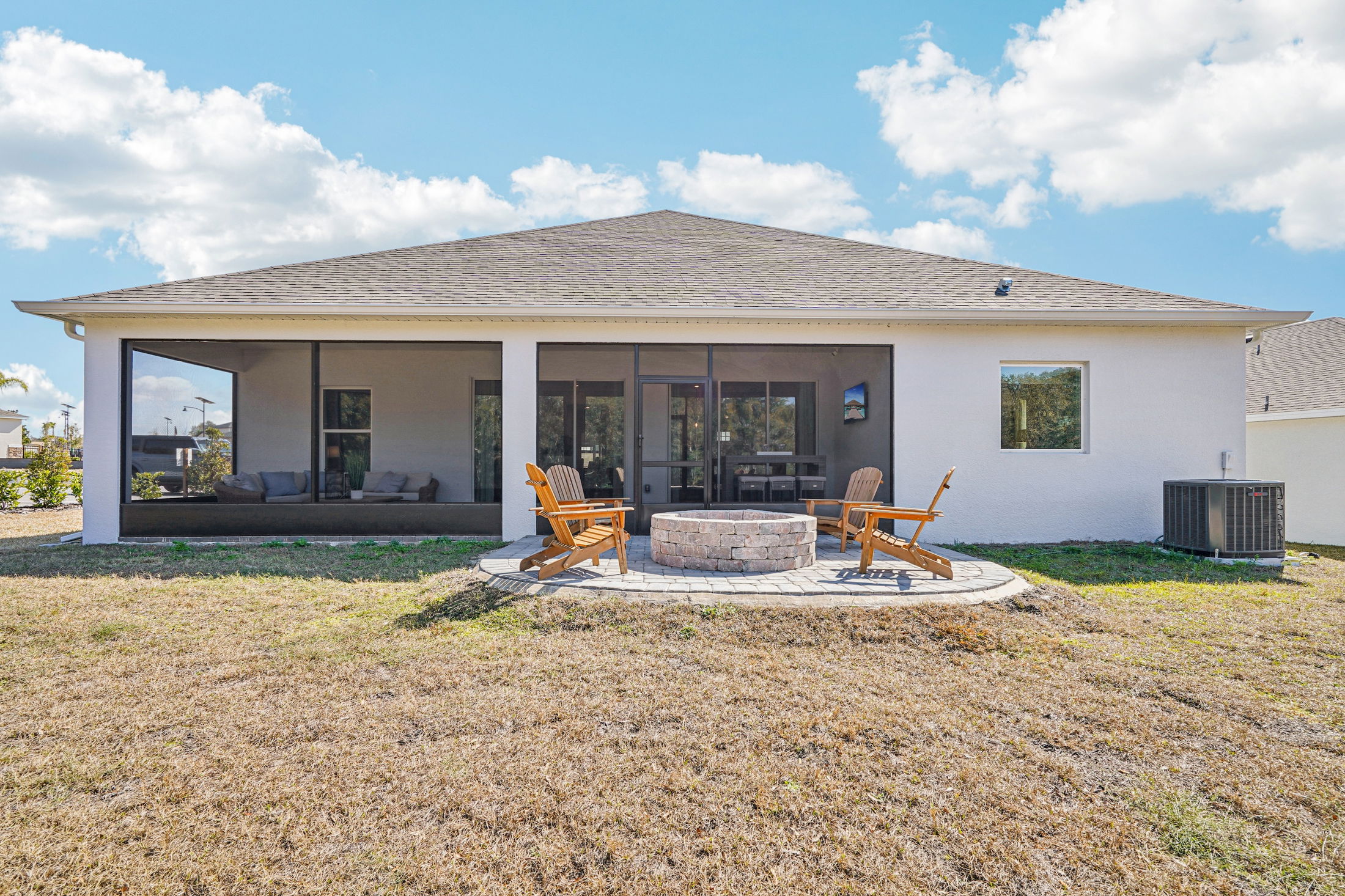 Modern backyard with a fire pit, wooden chairs, and a spacious screened porch under a blue sky with fluffy clouds.