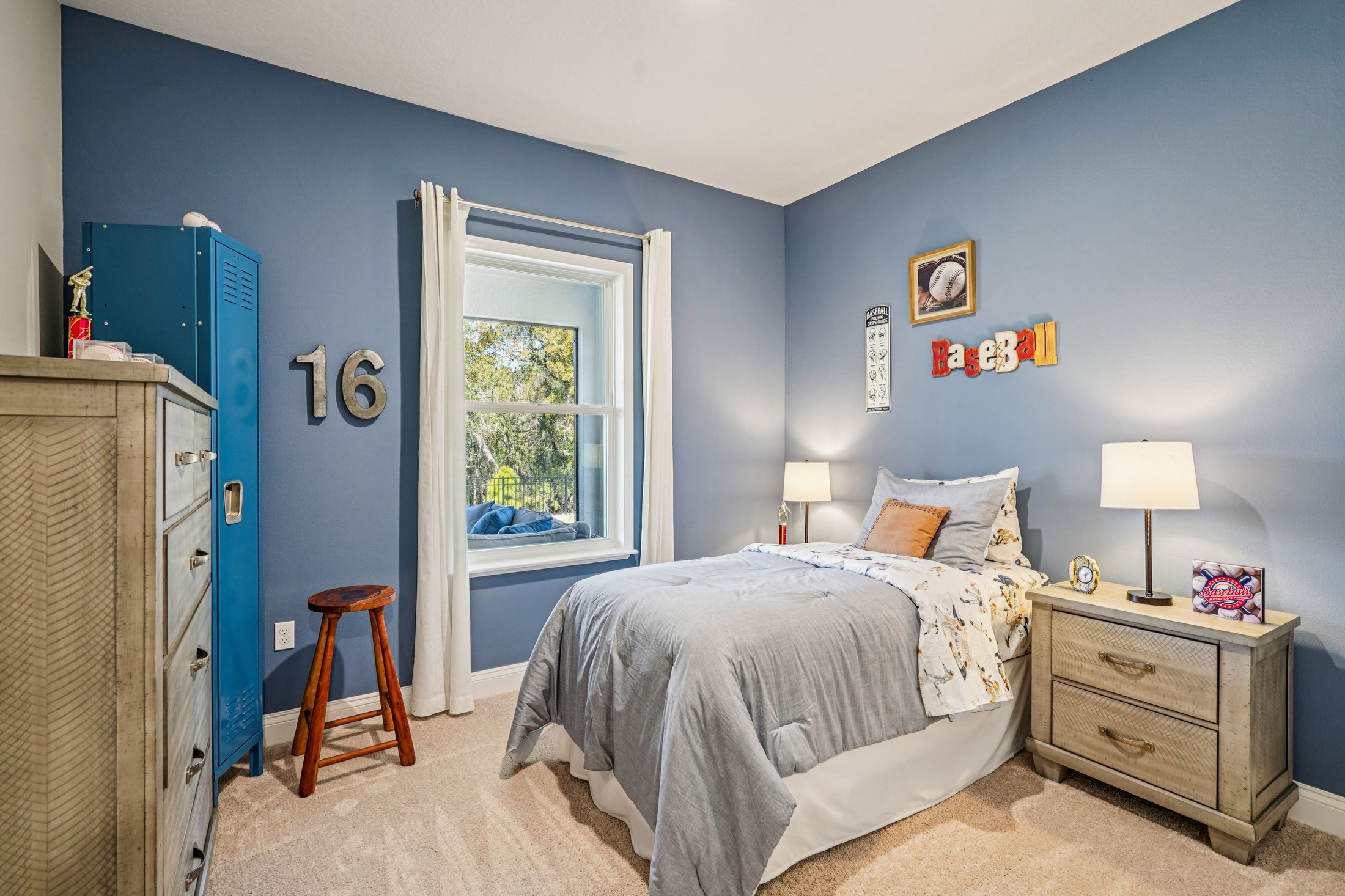 Cozy bedroom featuring blue walls, a twin bed with gray bedding, a wooden nightstand, a blue locker, and decorative baseball-themed items.
