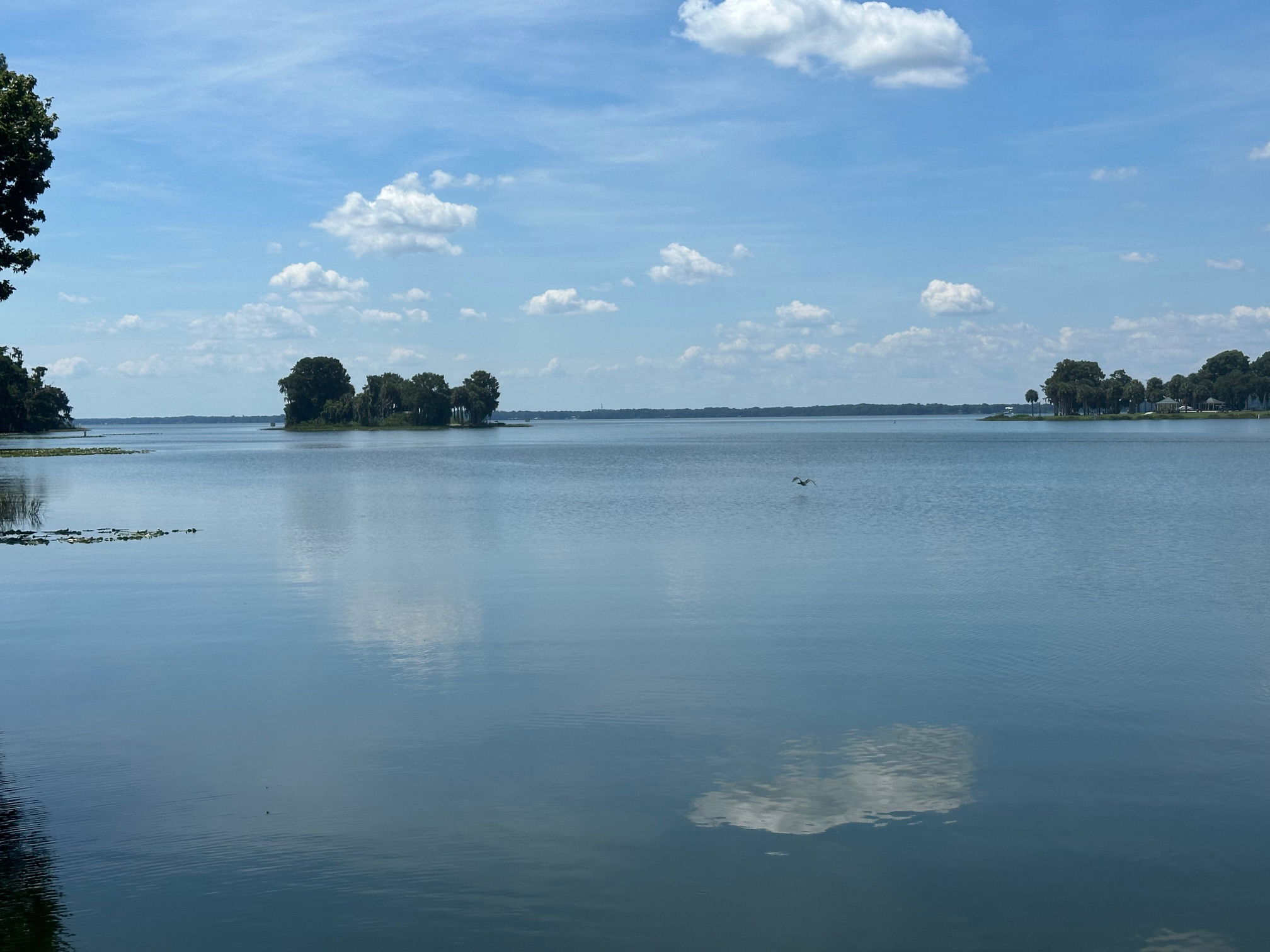 Serene lake view with small islands, calm blue water, and a partly cloudy sky reflecting on the surface.