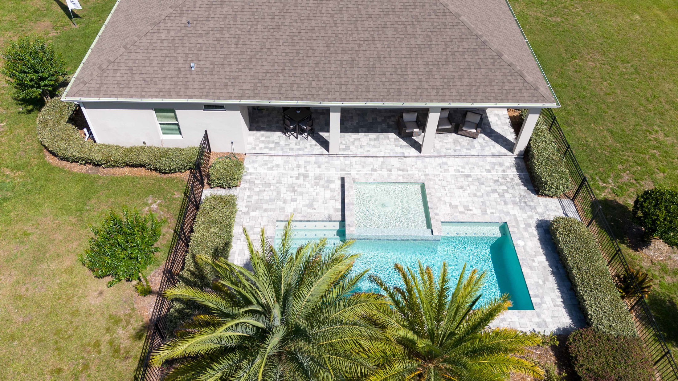 Aerial view of a modern backyard featuring a tiled patio, swimming pool with spa, and surrounding palm trees and greenery.