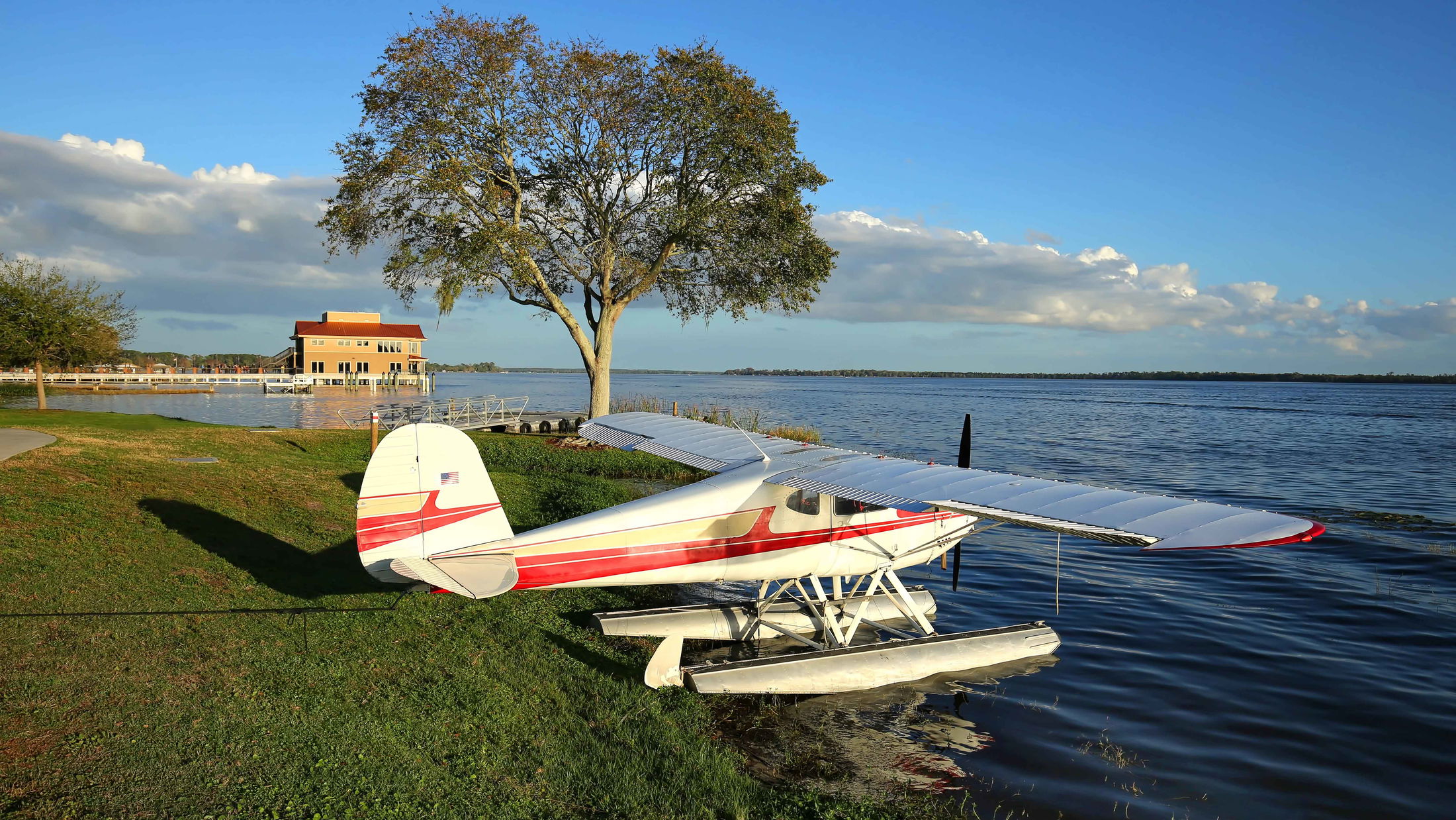 Seaplane parked by a serene lakeside with a tree and rustic building under a clear blue sky.