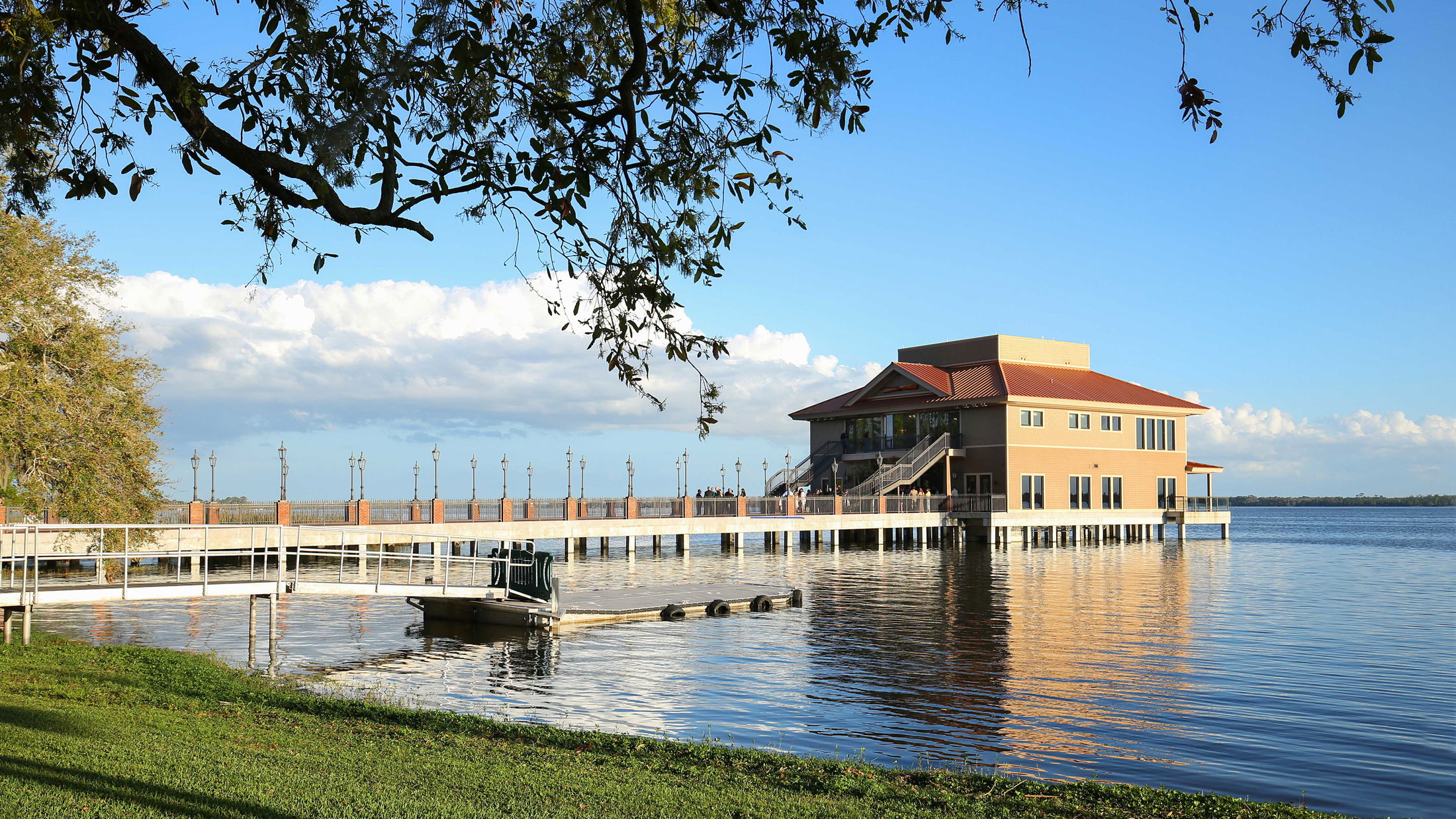 Waterfront building with a wooden dock and trees, reflected in calm lake waters on a clear, sunny day.