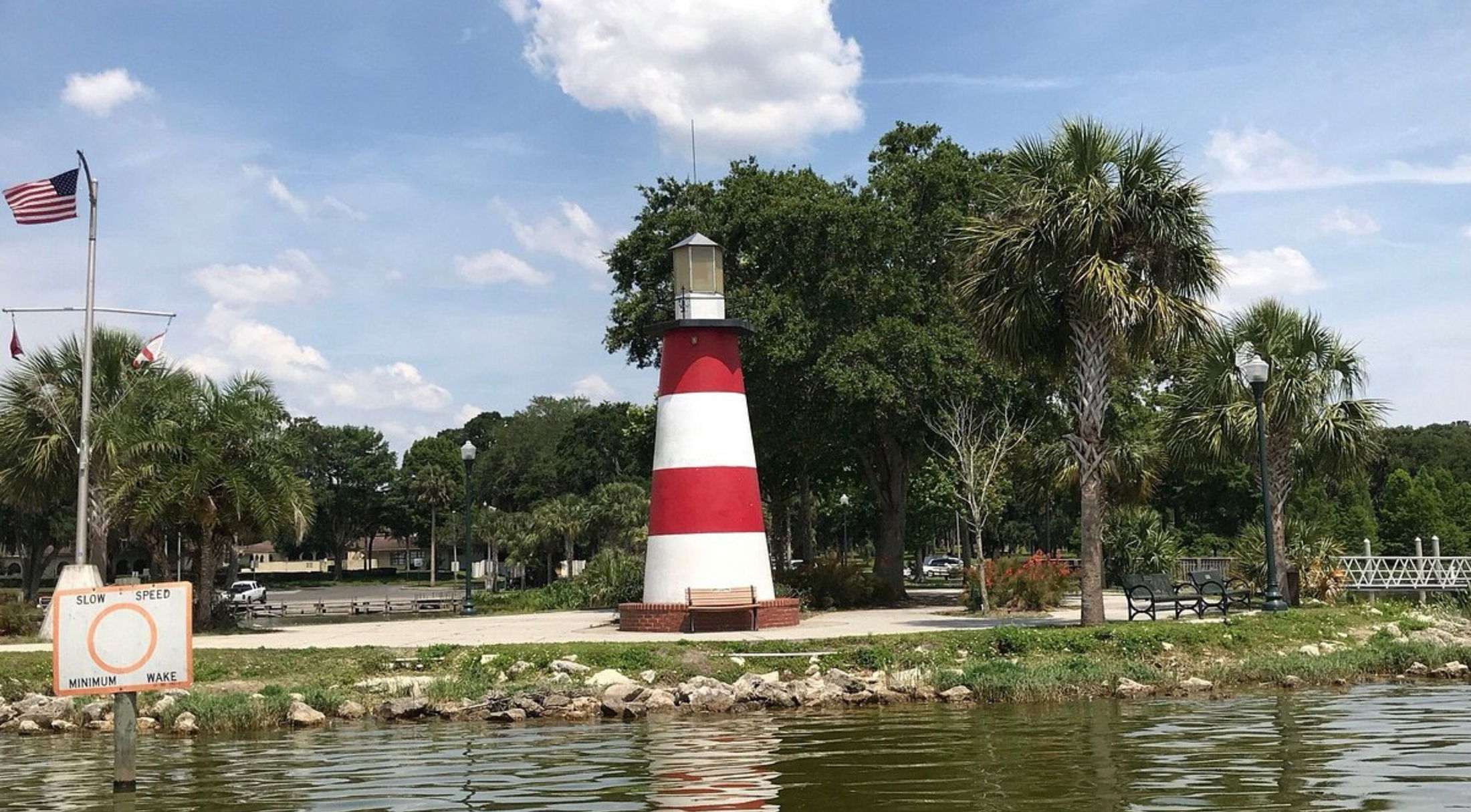Red and white striped lighthouse near a park with palm trees and a calm waterfront.