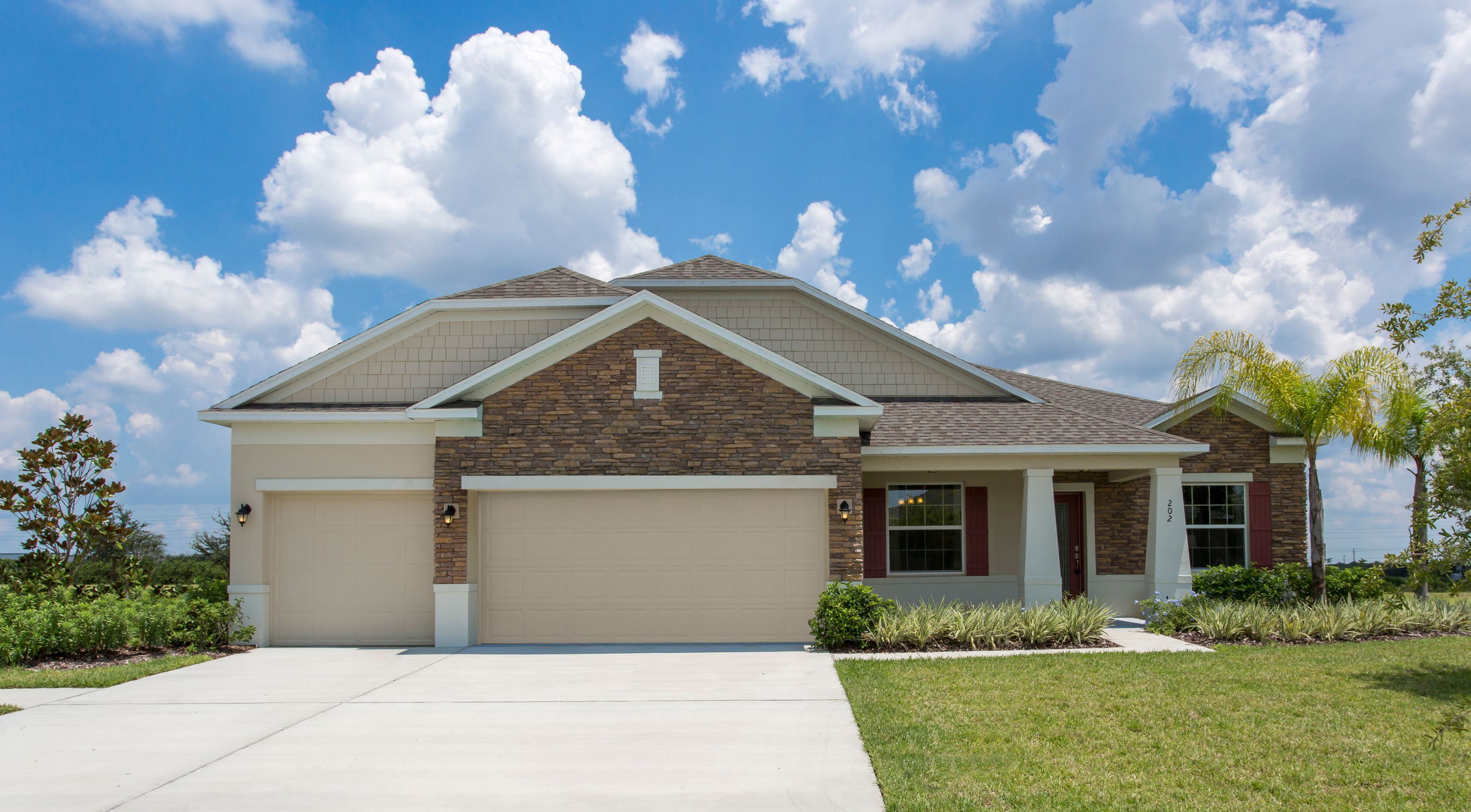 Modern suburban home with stone and siding facade, two-car garage, and well-manicured lawn under a blue sky with fluffy clouds.