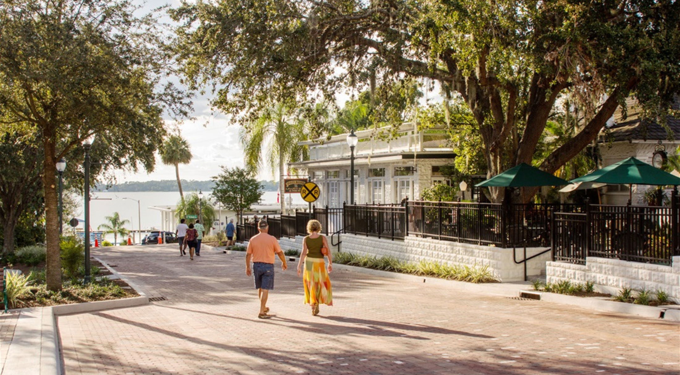 A couple walks along a picturesque tree-lined street in a coastal town, with charming shops, outdoor seating, and a view of the water.